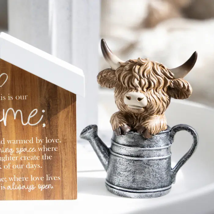 A decorative figurine of a shaggy brown highland cow sitting inside a galvanized metal watering can, displayed on a white surface next to a wooden house-shaped sign.