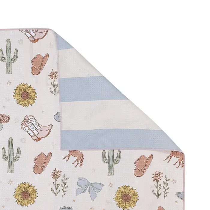 A folded fabric item, likely a blanket or towel, showing a western-themed print with sunflowers, cacti, cowboy boots, hats, and a horse on a cream background, with a reverse side featuring light blue and cream horizontal stripes.