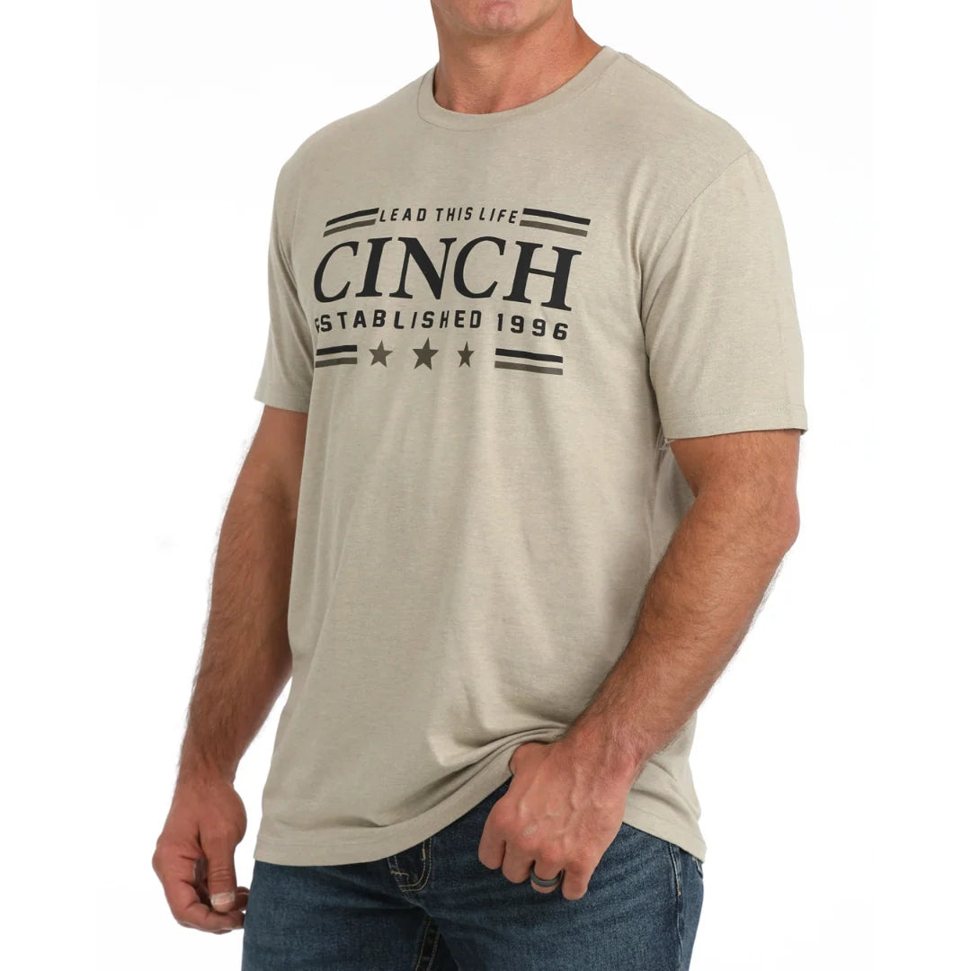 A man wearing a light beige crewneck t-shirt with a black graphic print that reads 'CINCH ESTABLISHED 1996'.