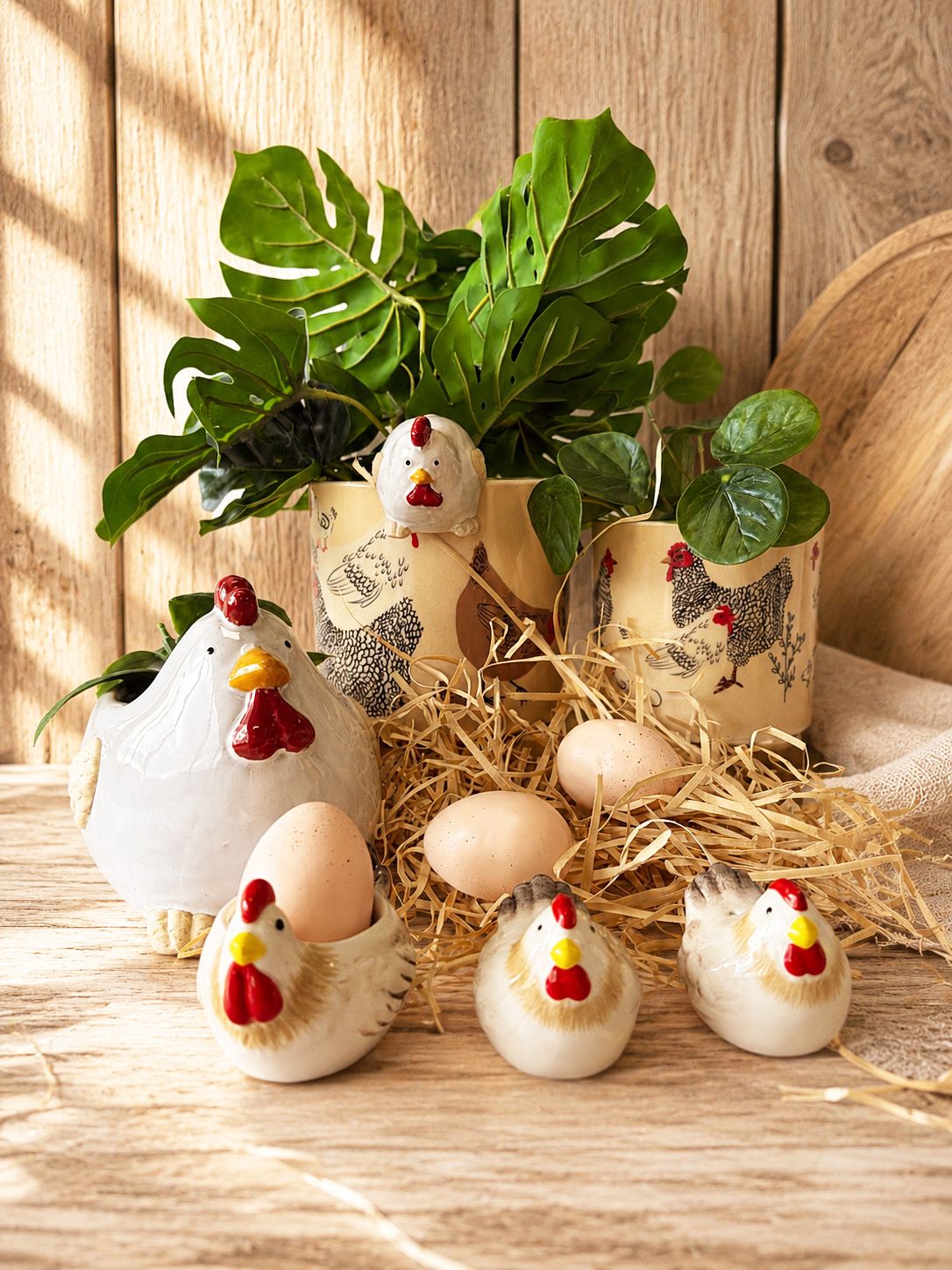 A rustic farm-themed decorative set featuring ceramic chicken figurines, potted plants in chicken-shaped pots, and faux eggs arranged on straw against a wooden plank background.