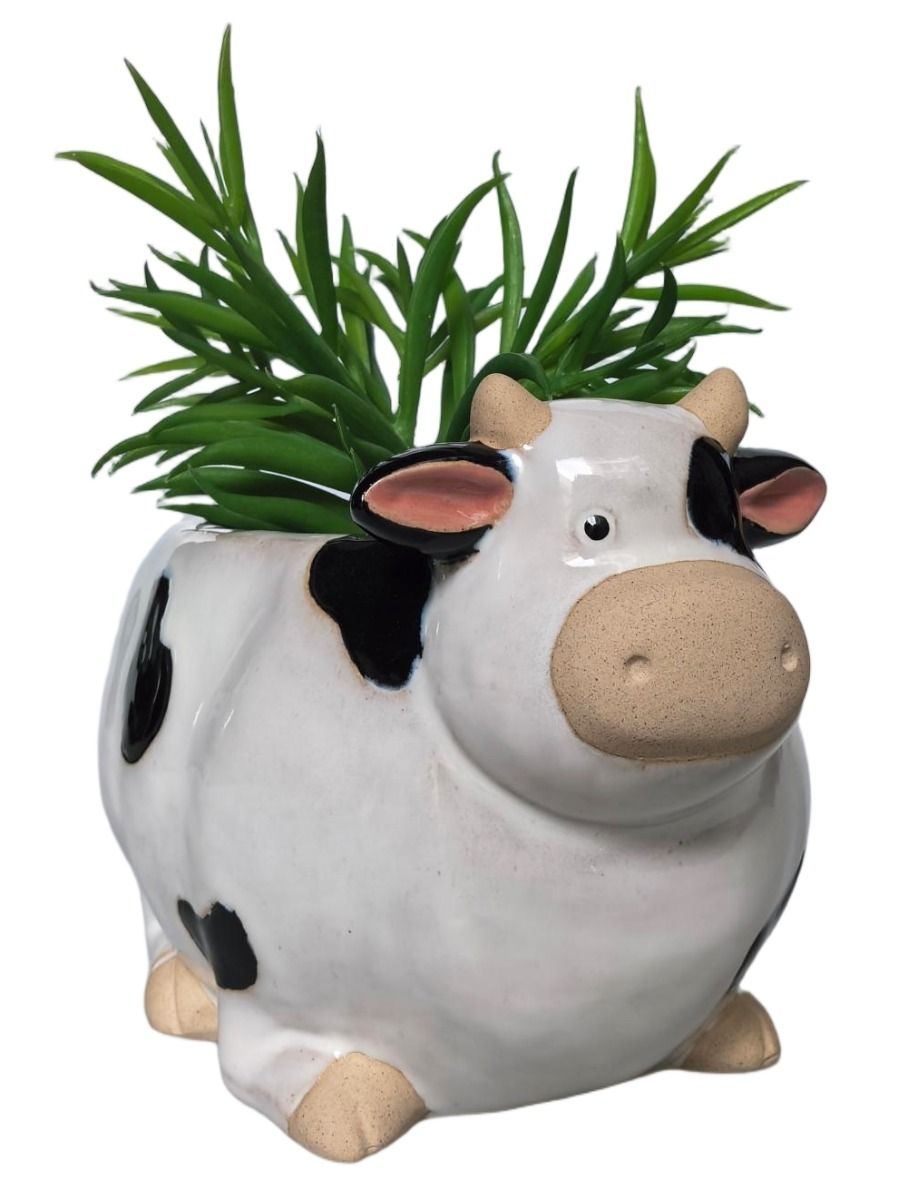 A ceramic cow-shaped planter with a glossy white body, black spots, pink inner ears, beige snout and hooves, and green artificial grass growing out of the top, shown on a white background.