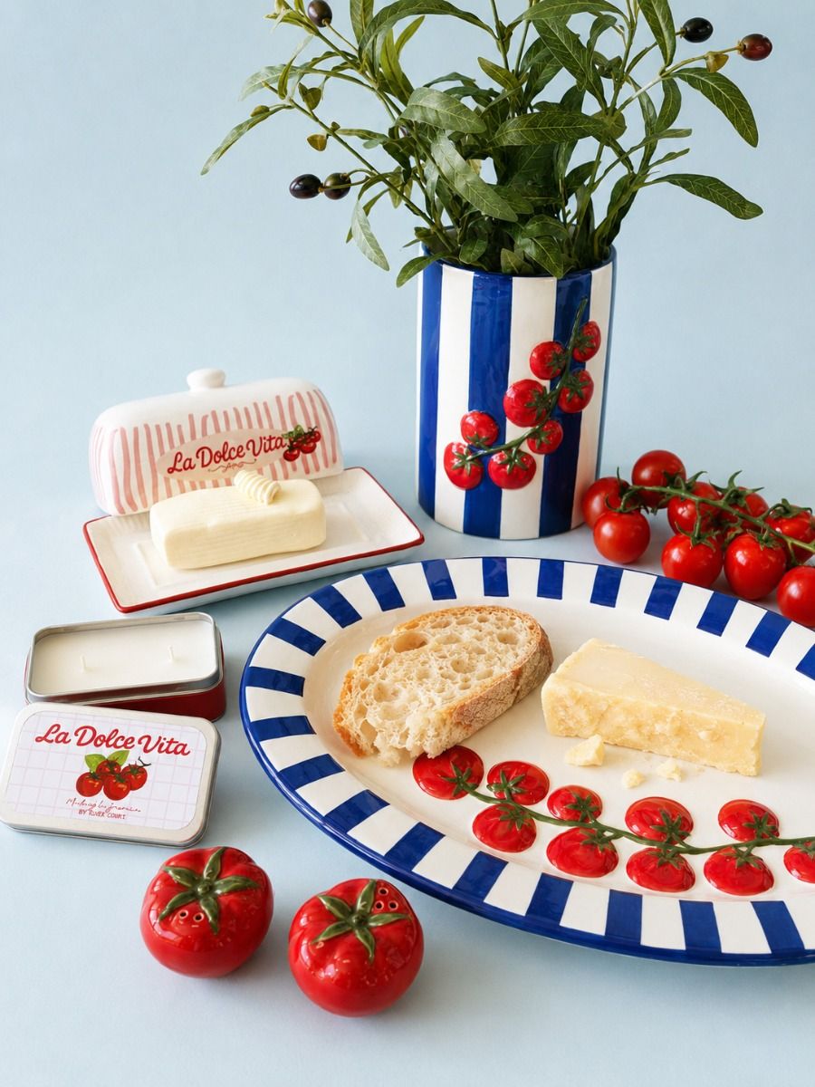 A styled tabletop scene with La Dolce Vita branded dishes and food props on a pale blue background. Visible products include a blue-and-white striped plate with red berry motifs, a matching butter dish, a small tin, and a vase with greenery; bread, cheese, and tomatoes are arranged as serving props.