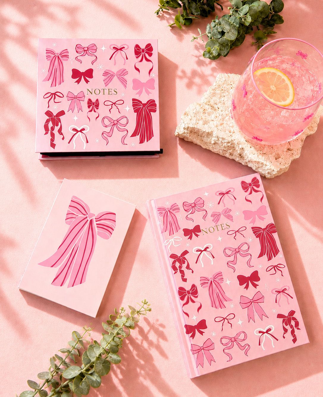 Pink notebook covers decorated with bow illustrations, arranged on a pink surface with a glass of pink drink, a stone slab, and greenery.
