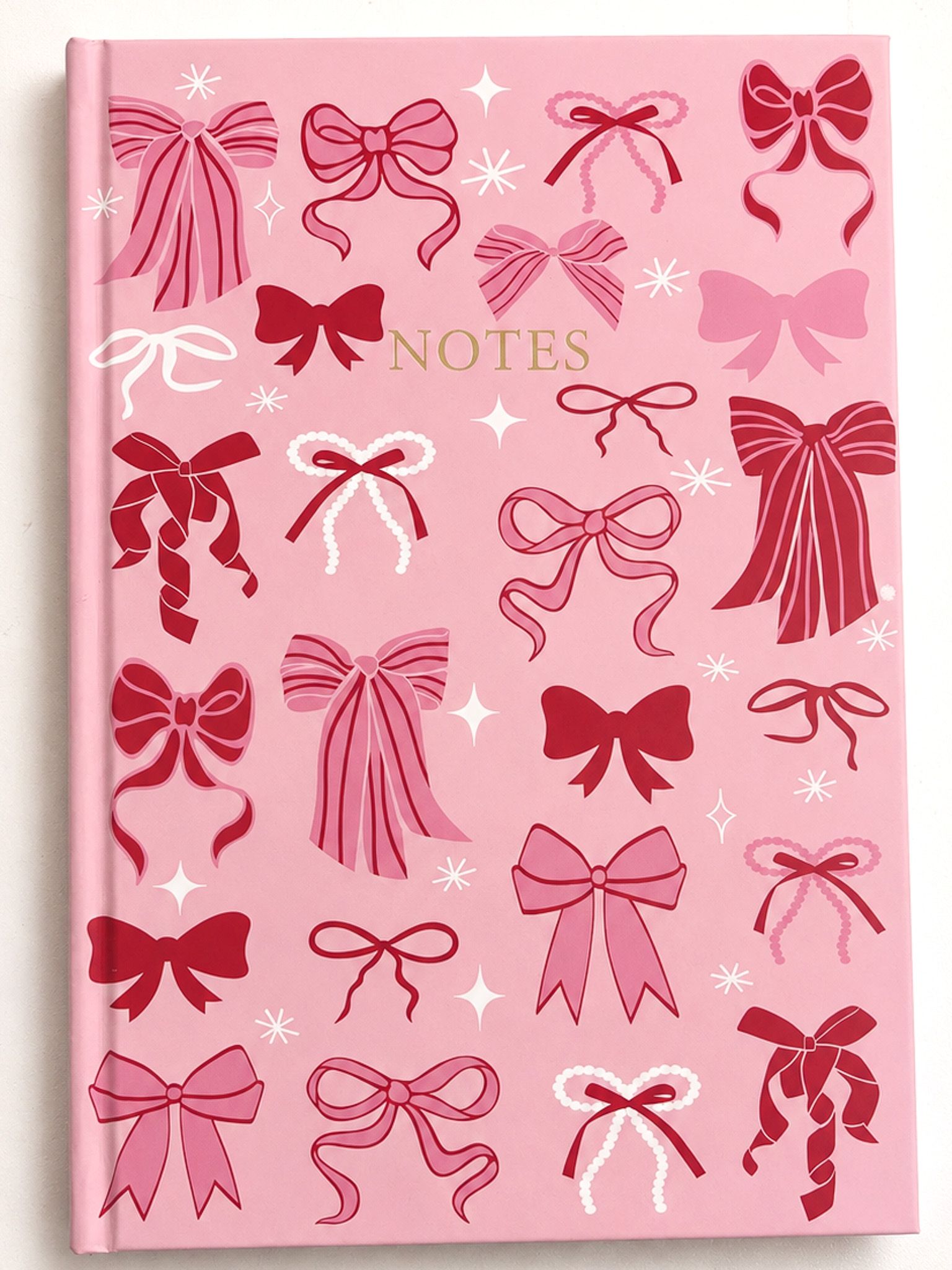 A pink hardcover notebook decorated with various red and pink bow illustrations and white star accents, featuring the word 'NOTES' in gold on the front.