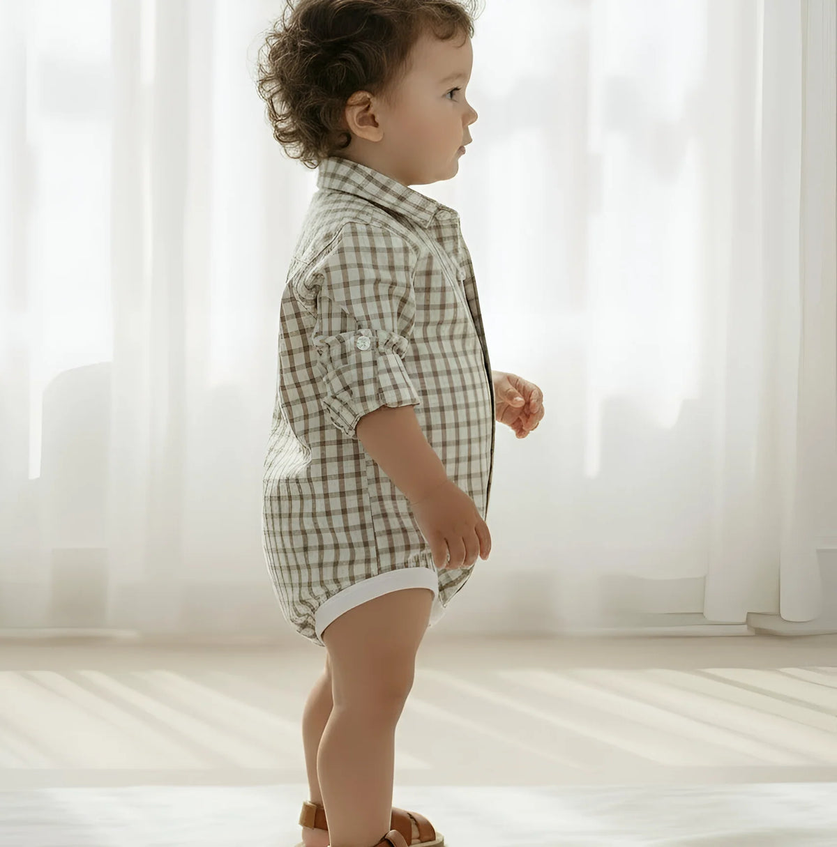 A toddler standing in profile wearing a light beige and brown gingham short-sleeve romper with white trim, photographed in a bright, softly lit room with sheer curtains.