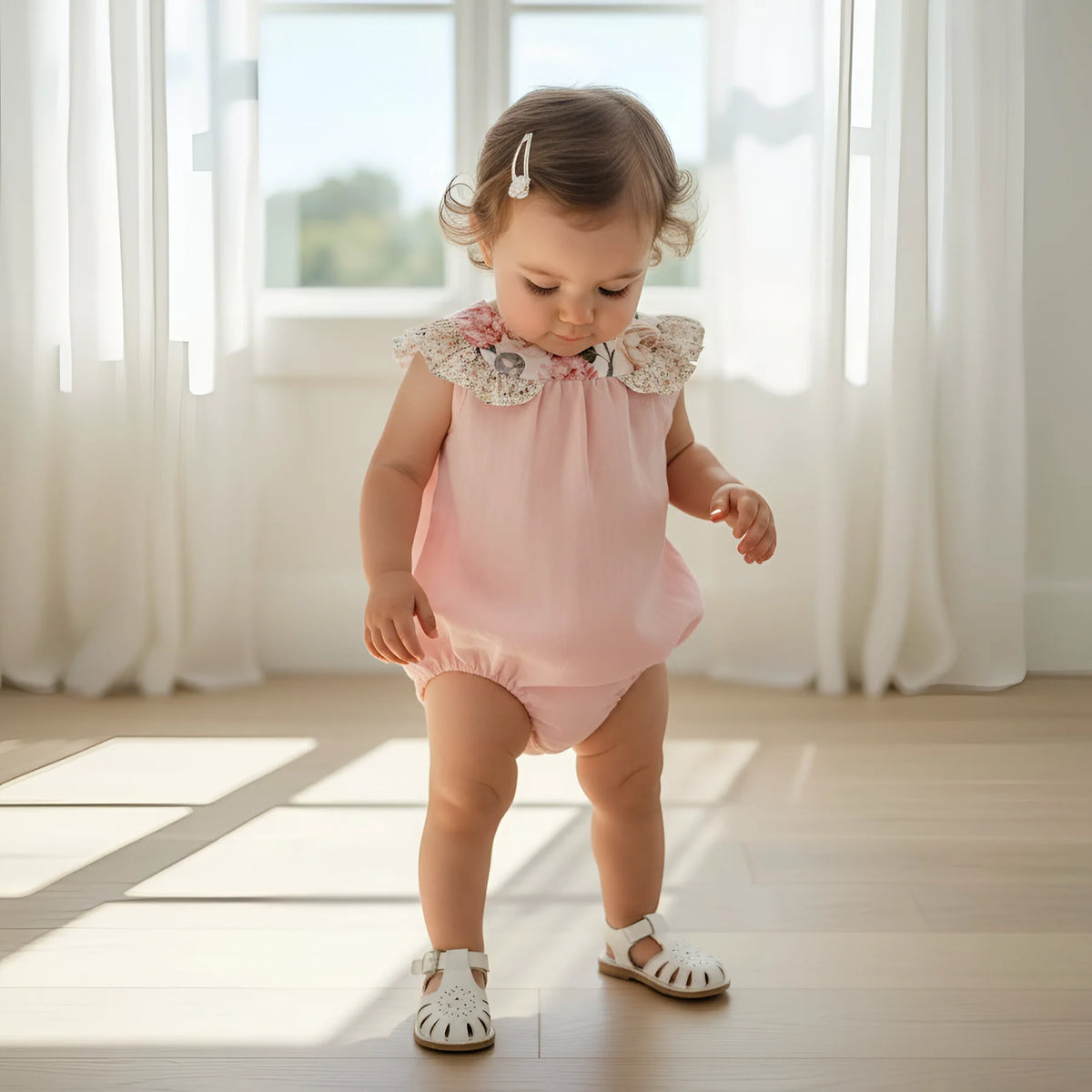 A toddler girl wearing a light pink sleeveless romper with a floral lace collar and white sandals, standing on a wooden floor in a bright room with white curtains.