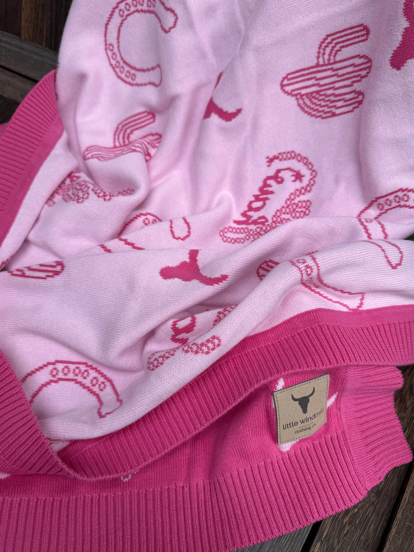 A close-up of a pink knit blanket featuring a darker pink cactus and snake print, finished with a bright pink ribbed trim and a small tan brand patch.