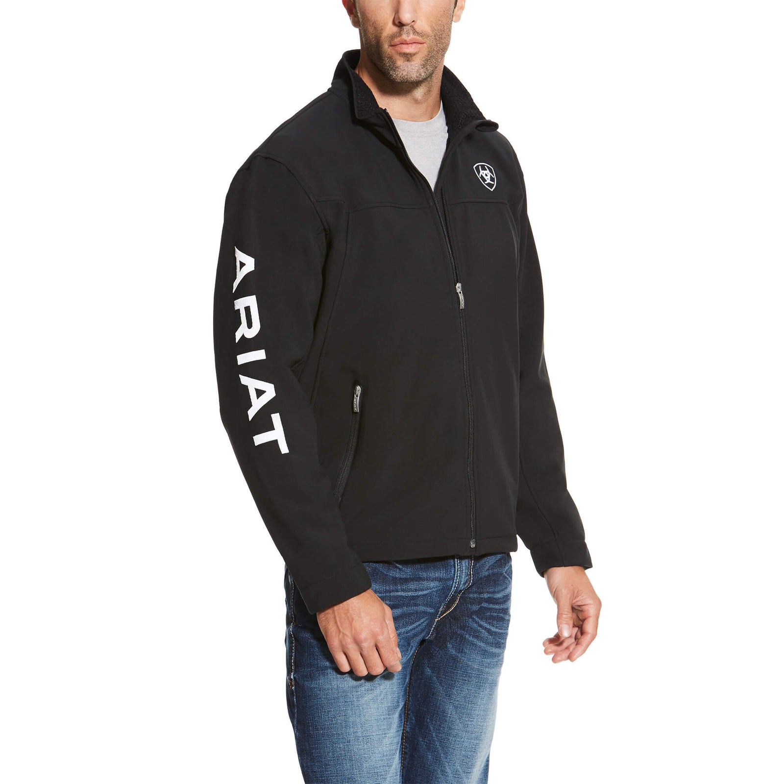 Ariat Men's Softshell Team Jacket. A man wearing a black softshell team jacket with the brand name 'ARIAT' written across the sleeve.