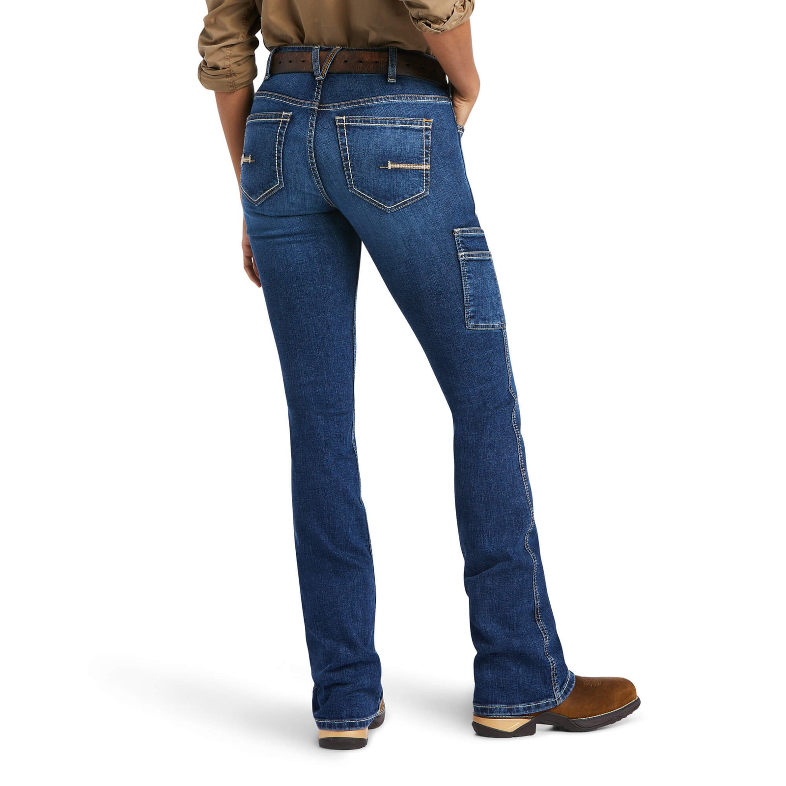 A woman wearing Ariat women's rebar flex perfect rise jeans in a blue denim color, standing with her hands in the pockets.