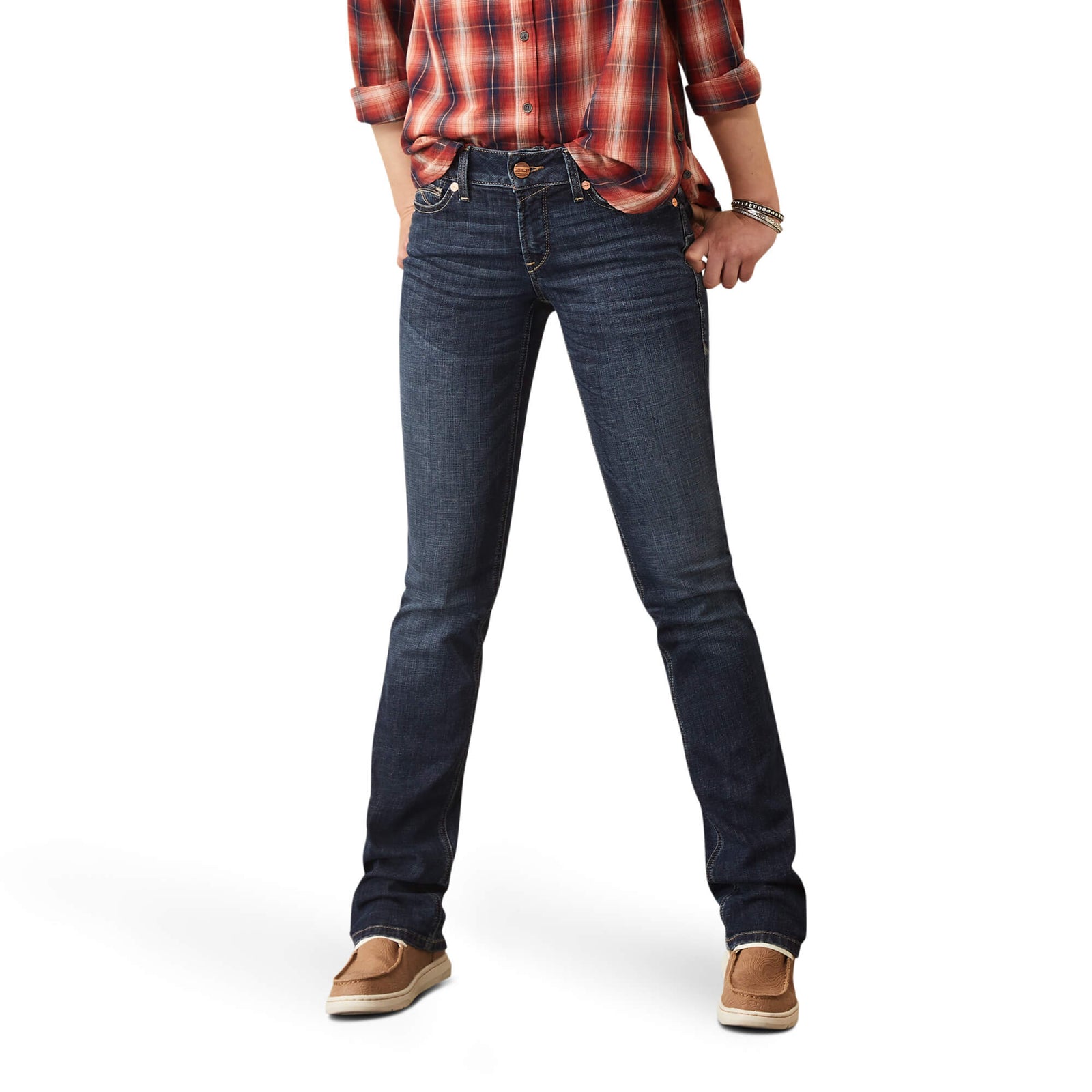 A woman standing, wearing Ariat Women's Aubree Perfect Rise Jean with a subtle 'A' design on the back pocket, a plaid shirt, and sneakers.