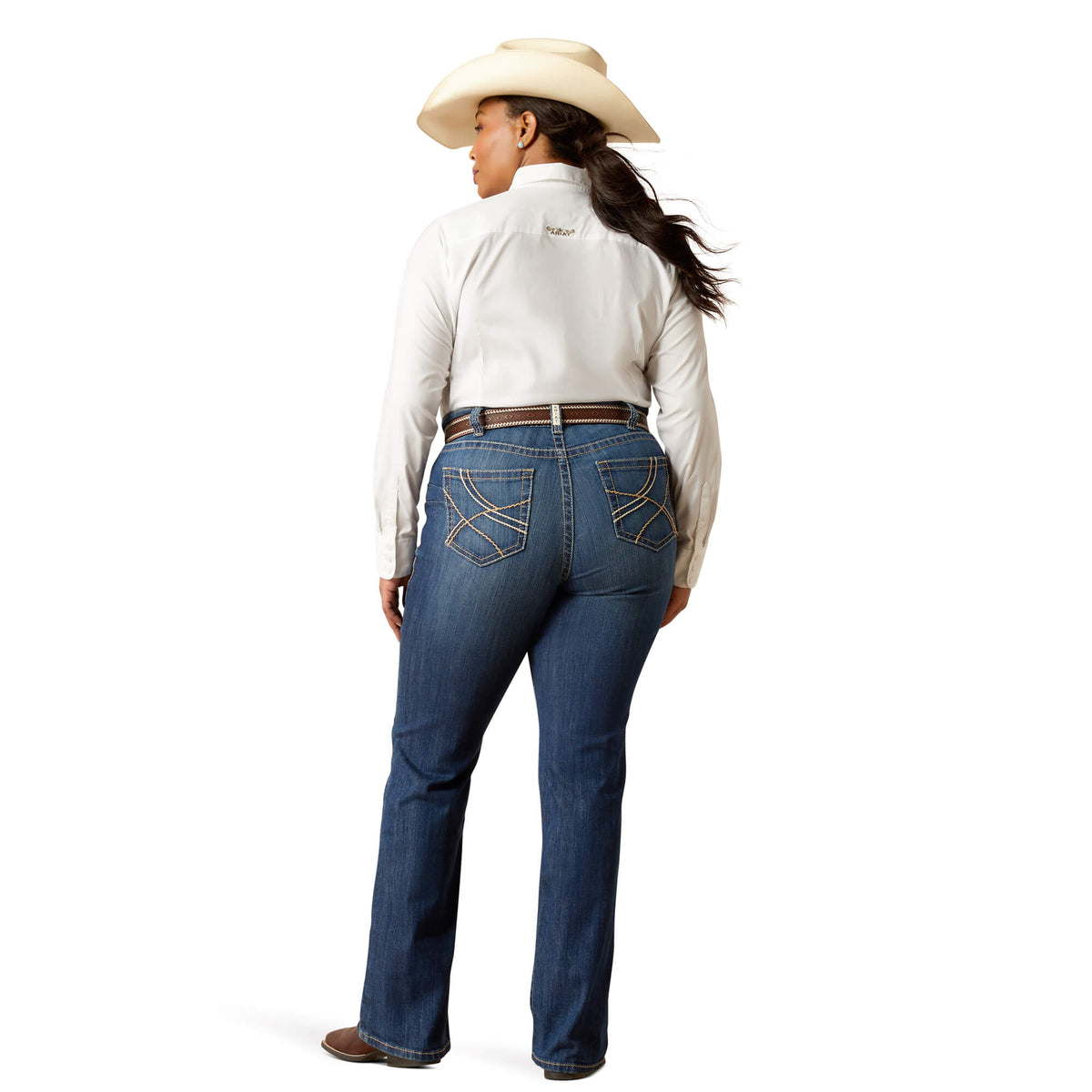 A woman wearing Ariat's women's Everlee Mid Rise Plus Size Jean, a straight leg denim jean, in a blue color. She is paired with a white shirt and a hat, standing with her back towards the camera.