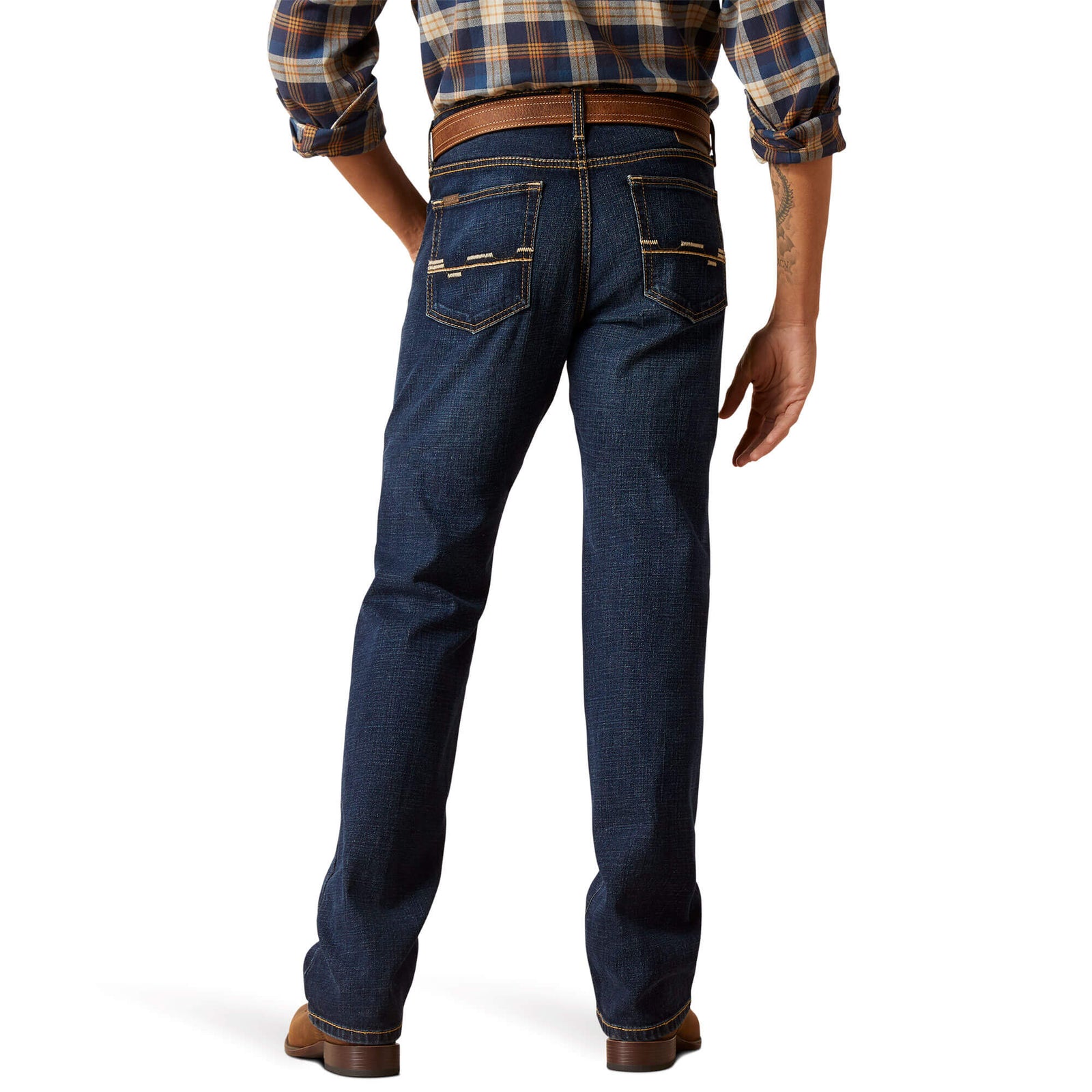 A man standing wearing Ariat Men's M8 Reese Modern Fit Jeans and brown boots. The jeans are blue with a subtle pattern, and the man has his hands in his pockets.