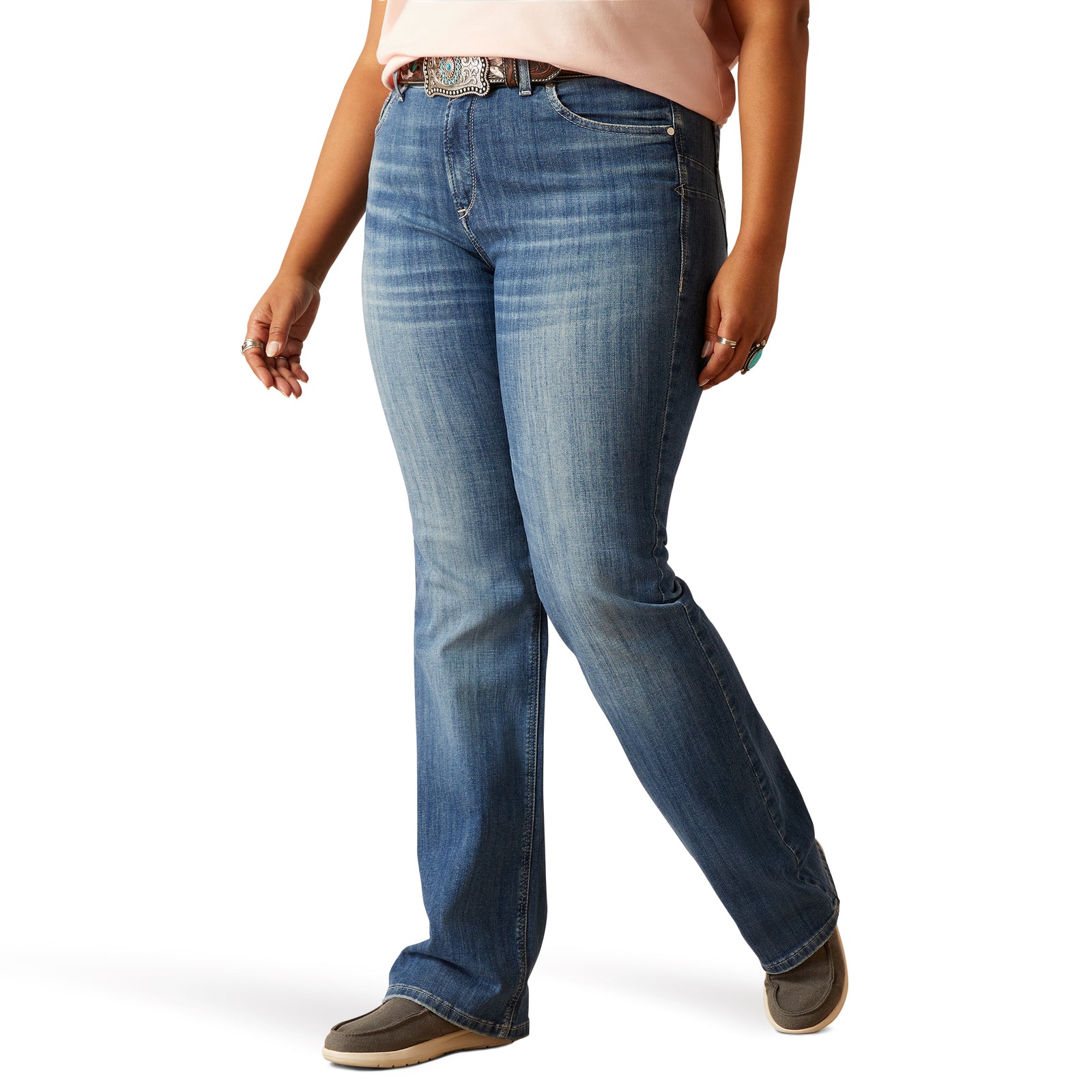 Ariat Women's Annie Perfect Rise Boot Cut Jean. A woman standing side-on wearing Ariat women's boot-cut jeans in a medium wash with raw-edge details on the back pocket.