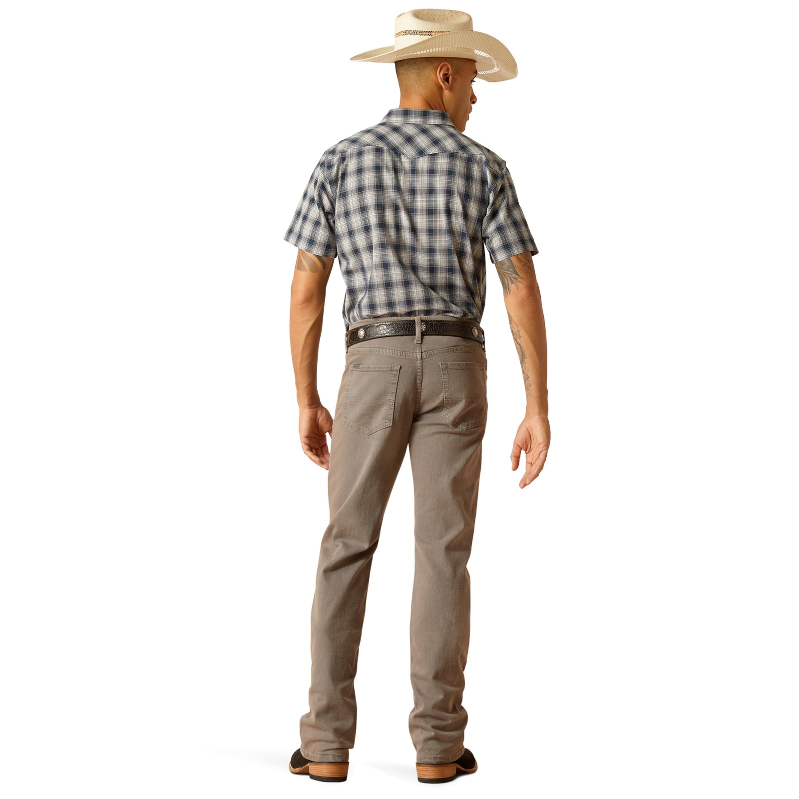 Ariat Men's M5 Hansen Straight Leg Jean. A man standing straight, wearing brown straight leg jeans and a checkered shirt with the sleeves rolled up. He has a belt with a prominent buckle and is feet are placed shoulder-width apart.
