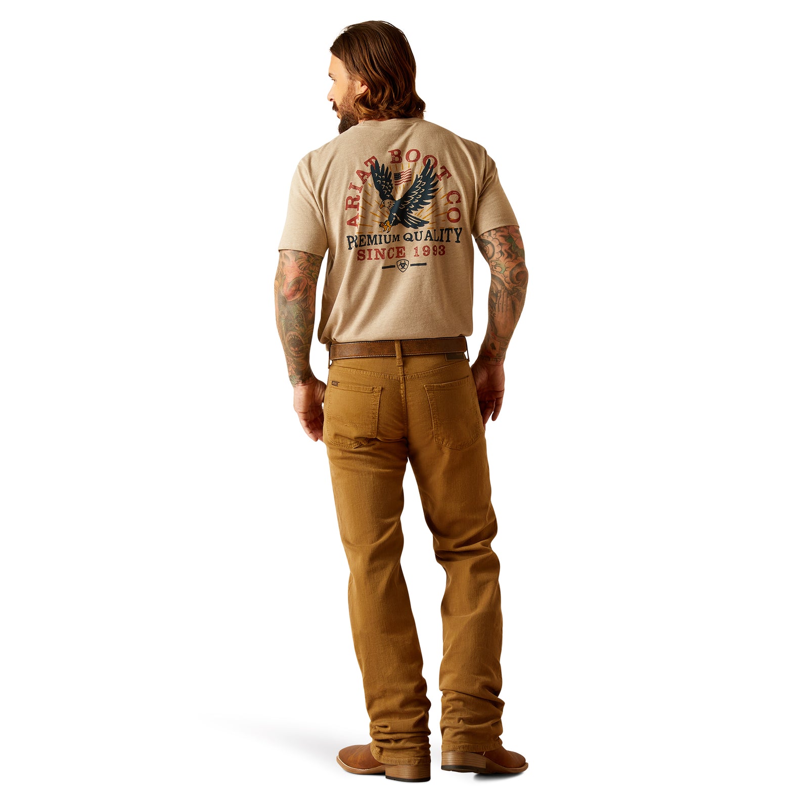 Ariat Men's M5 Hansen Straight Leg Jean. A man wearing brown straight leg jeans and brown cowboy boots, with a visible tattoo on the upper left arm.