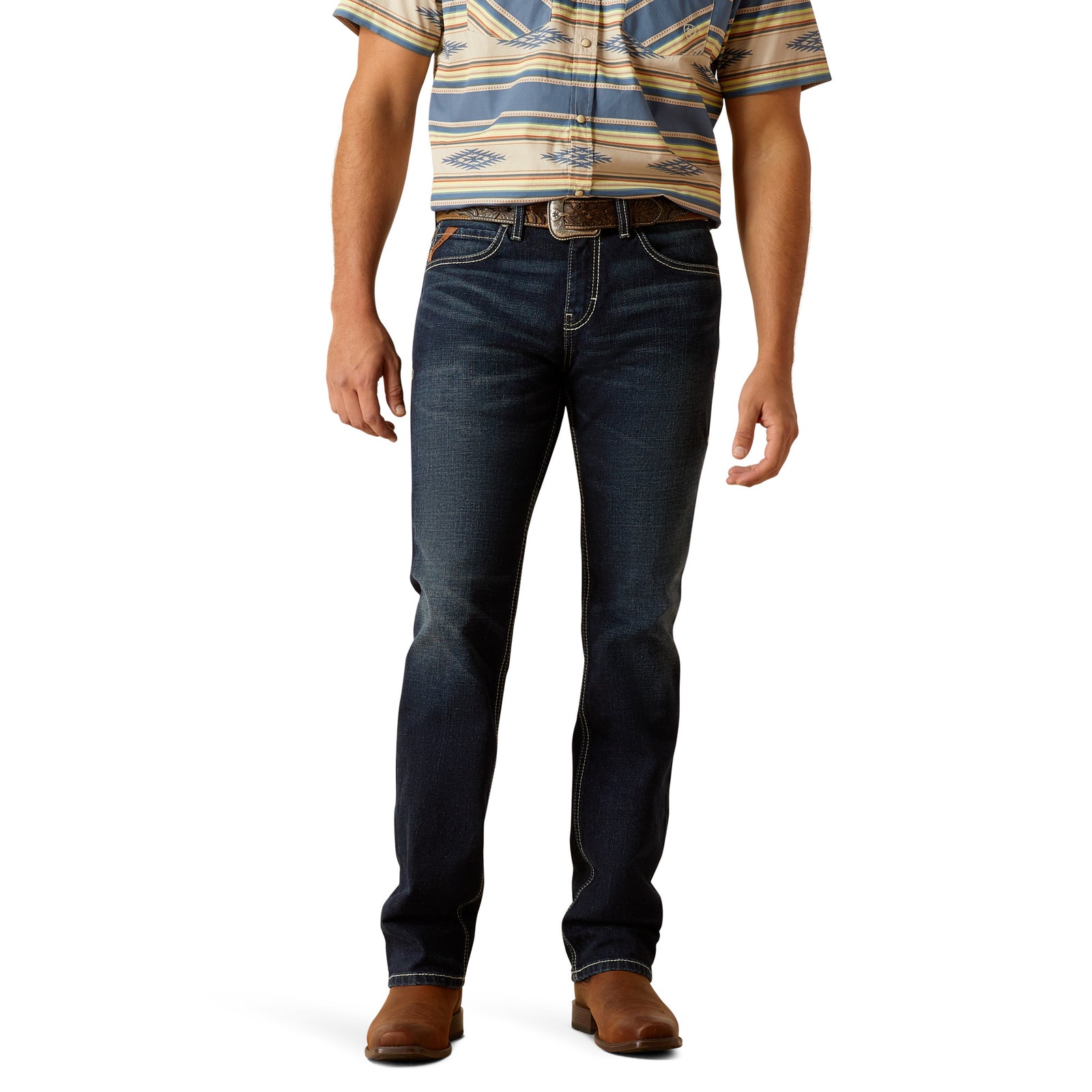 A man standing wearing Ariat M8 Antelope Men's Modern Slim Leg Jean, a belt, and a striped shirt with brown boots.