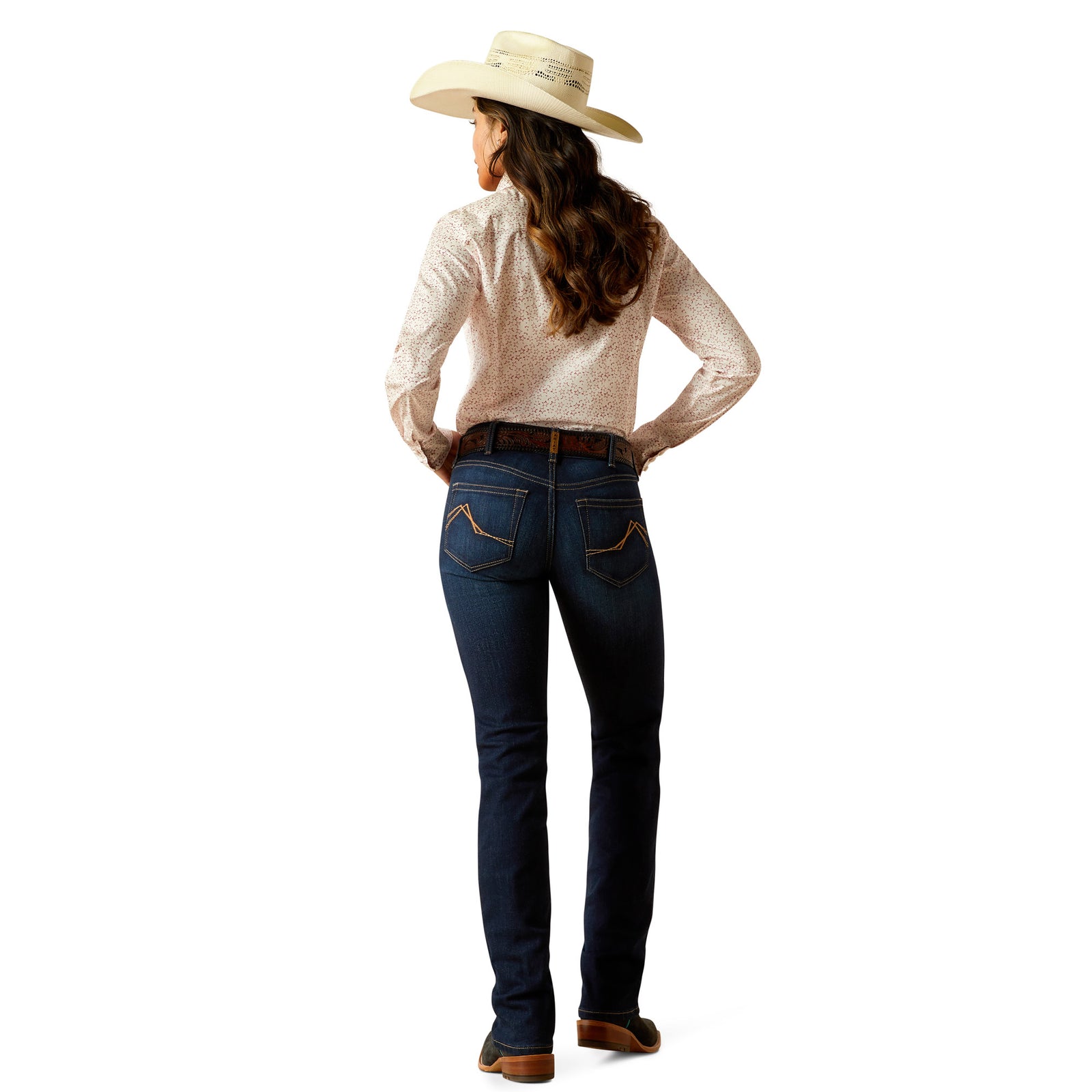 Ariat Women's Romana Mid Rise Boot Cut Jean. A woman standing, wearing mid-rise boot cut jeans with copper stitching, a light colored top, and brown boots.