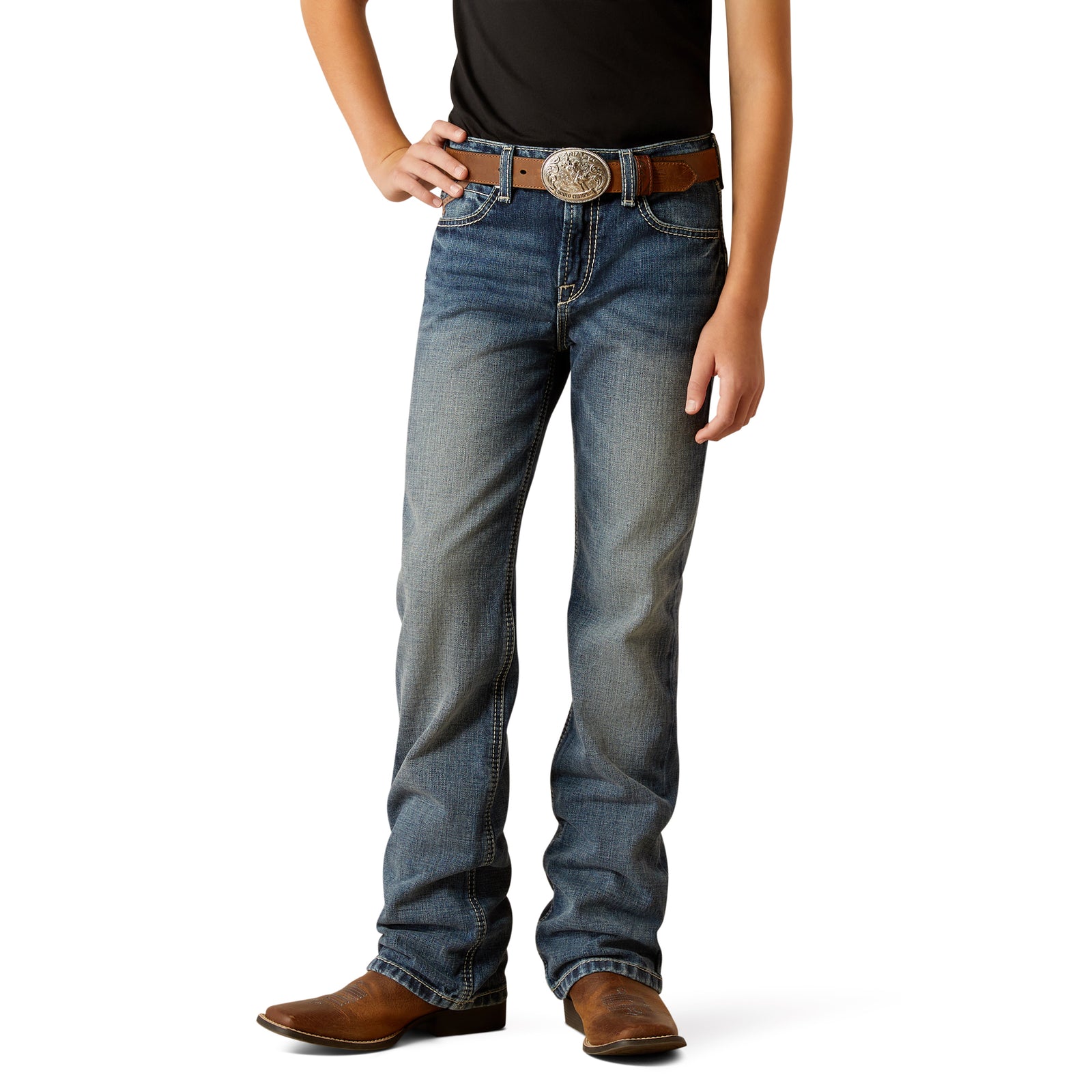 Ariat Boy's B4 Challenger Boot Cut Jean. A boy standing, wearing a pair of Ariat boot cut jeans and brown leather boots, with a belt and belt buckle visible.