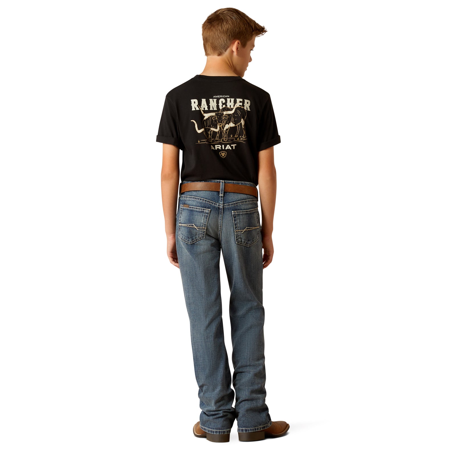 Ariat Boy's B4 Challenger Boot Cut Jean. A boy standing, wearing a pair of Ariat boot cut jeans and brown leather boots, with a belt and belt buckle visible.