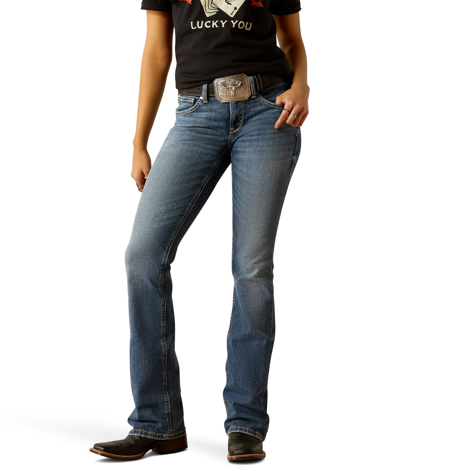 A woman standing, wearing Ariat Women's Perfect Rise Brittany Boot Cut Jean with a light-medium wash and a belt with a silver buckle.