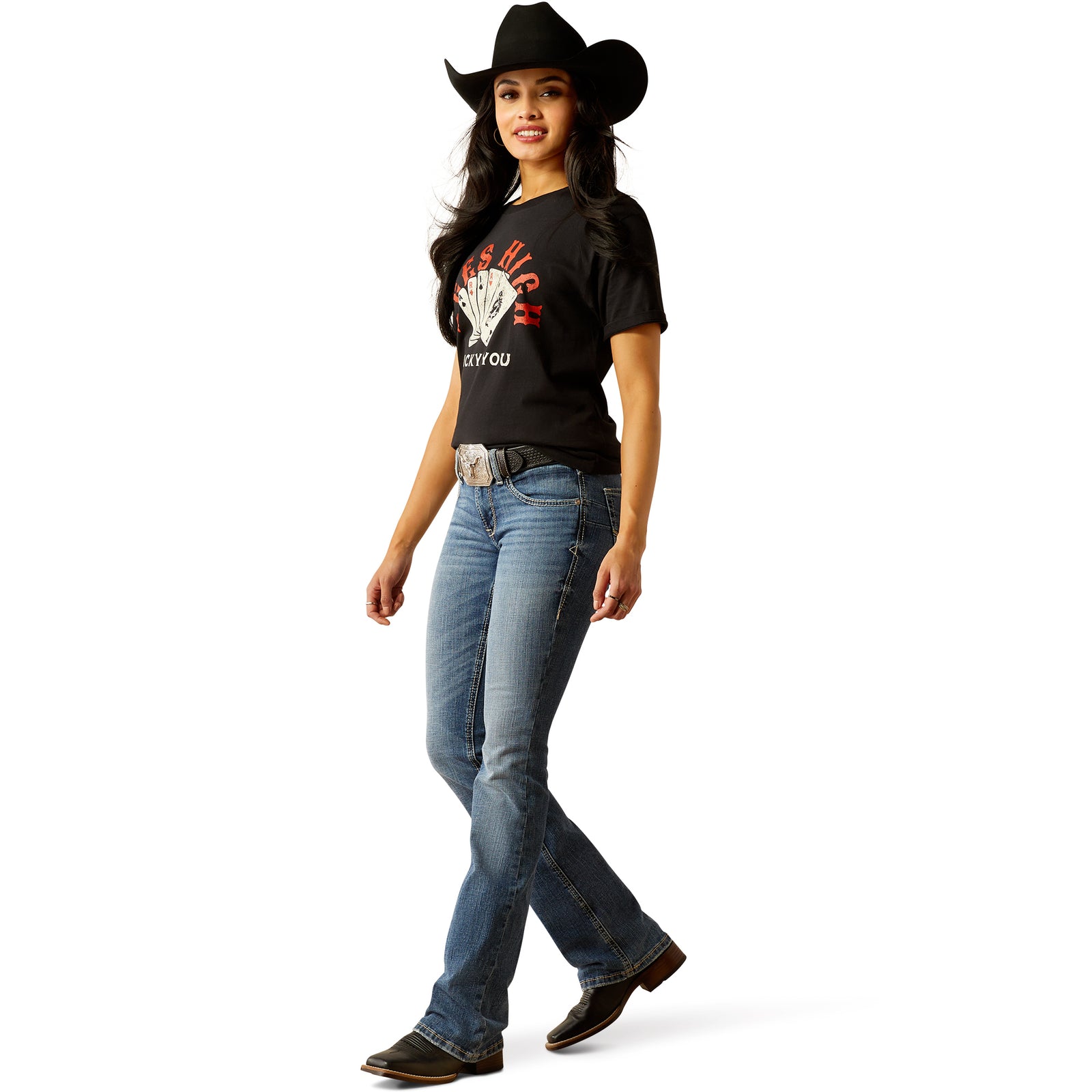 A woman standing, wearing Ariat Women's Perfect Rise Brittany Boot Cut Jean with a light-medium wash and a belt with a silver buckle.