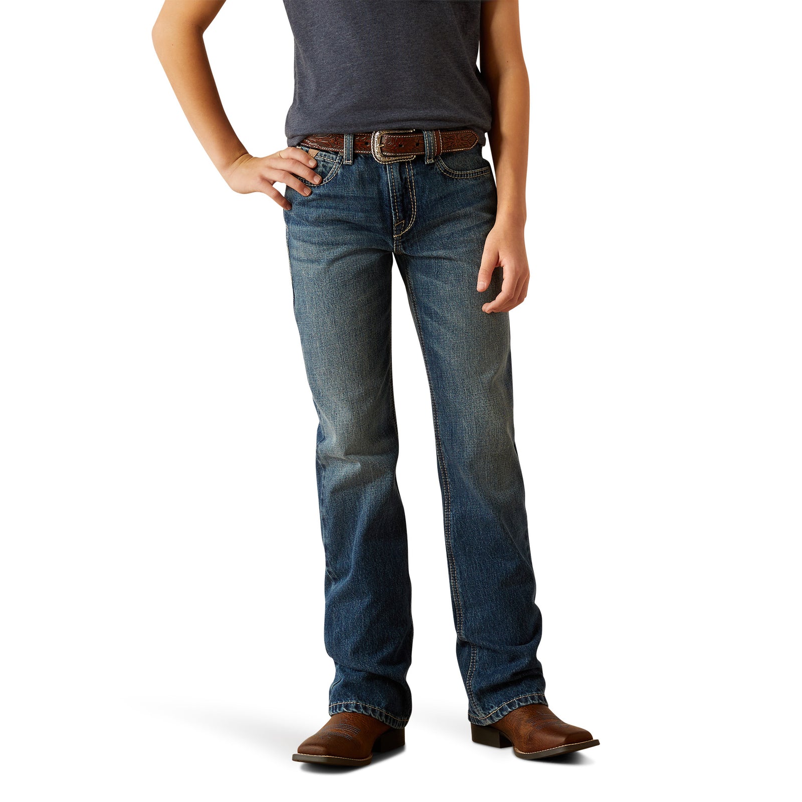 Ariat Boy's B4 Relaxed Kelvin Boot Cut Jean. A boy standing with one hand on his hip, wearing a pair of Ariat boy's relaxed fit boot cut jeans and a belt, with a pair of boots.