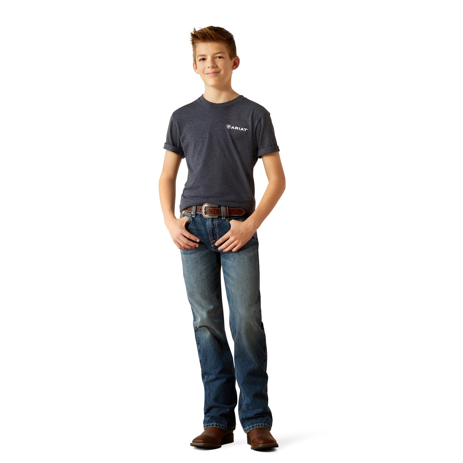 Ariat Boy's B4 Relaxed Kelvin Boot Cut Jean. A boy standing with one hand on his hip, wearing a pair of Ariat boy's relaxed fit boot cut jeans and a belt, with a pair of boots.