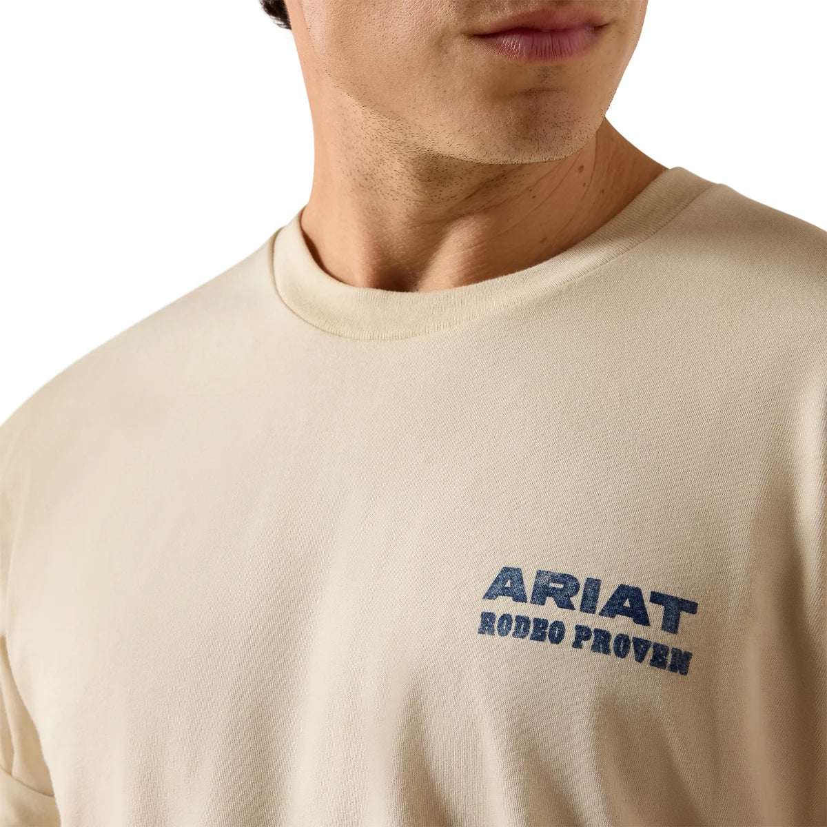 Close-up of a person wearing a cream-colored crewneck t-shirt with a dark blue 'ARIAT RODEO PROVEN' logo printed on the left chest.