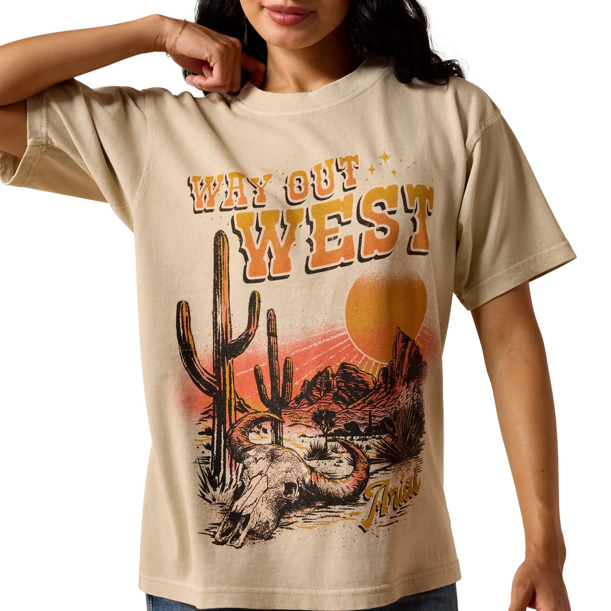 A woman modeling a beige graphic t-shirt with a western design that reads 'WHY GO WEST' above a desert landscape and skull graphic, paired with denim shorts against a white background.