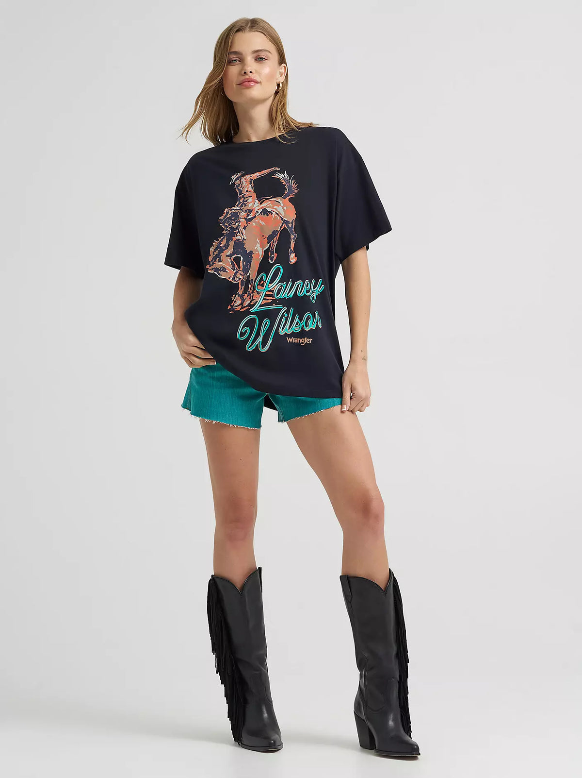 Woman wearing a black t-shirt with a graphic design and text, turquoise shorts, and black boots on a white background.
