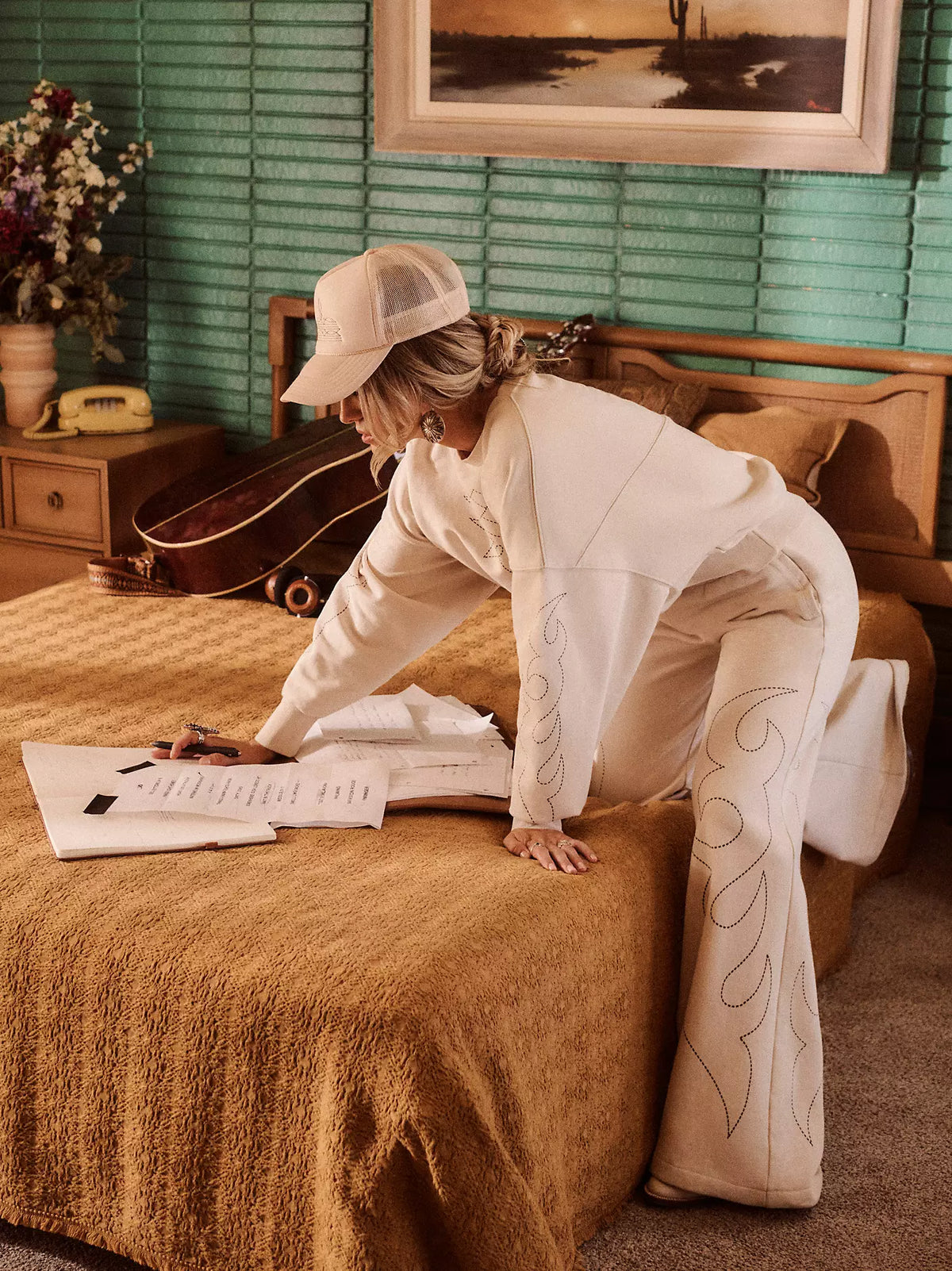 Person in a white outfit with decorative patterns on pants, sitting on a brown couch in a room with green walls and a guitar.