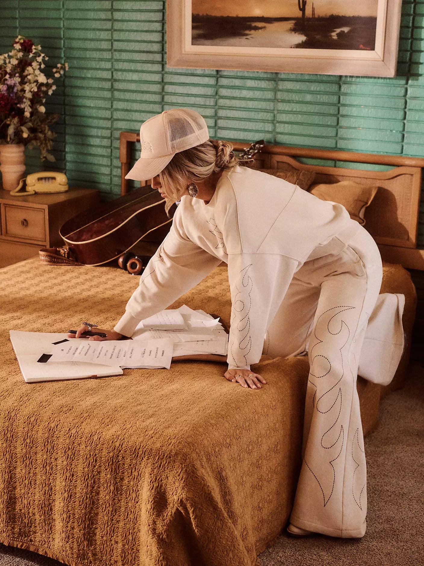Person in a white outfit with decorative patterns on pants, sitting on a brown couch in a room with green walls and a guitar.