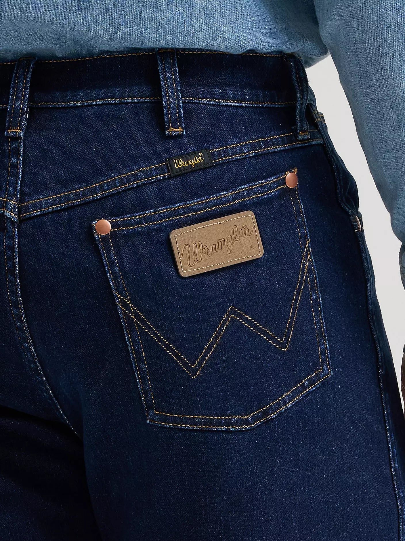 Close-up of the back pocket of dark blue denim jeans with a tan leather Wrangler patch and contrast stitching, worn with a light blue denim shirt.
