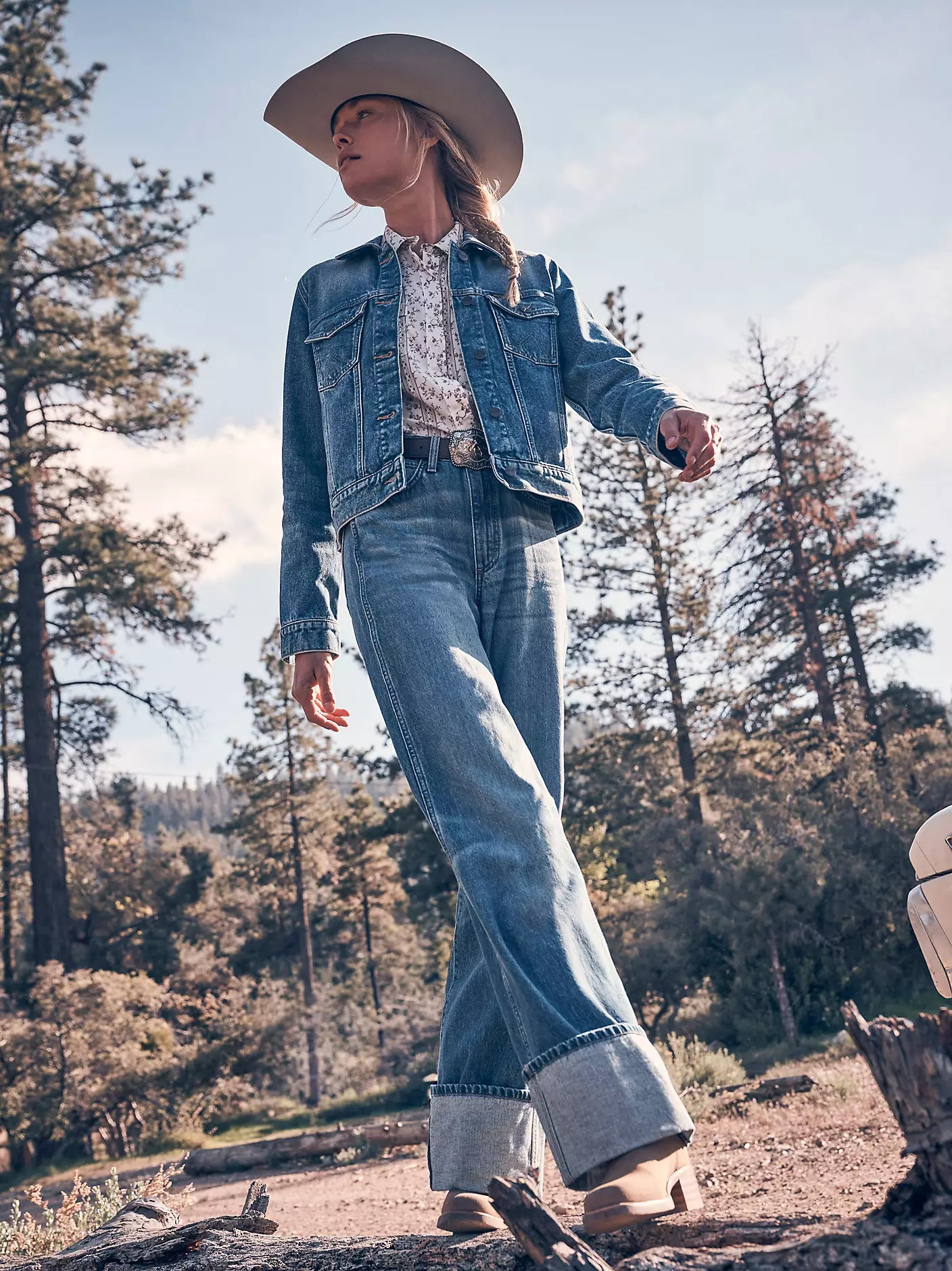 A woman wearing a light brown cowboy hat, a blue denim jacket, a white patterned shirt, and matching wide-leg blue jeans stands outdoors in a wooded setting.
