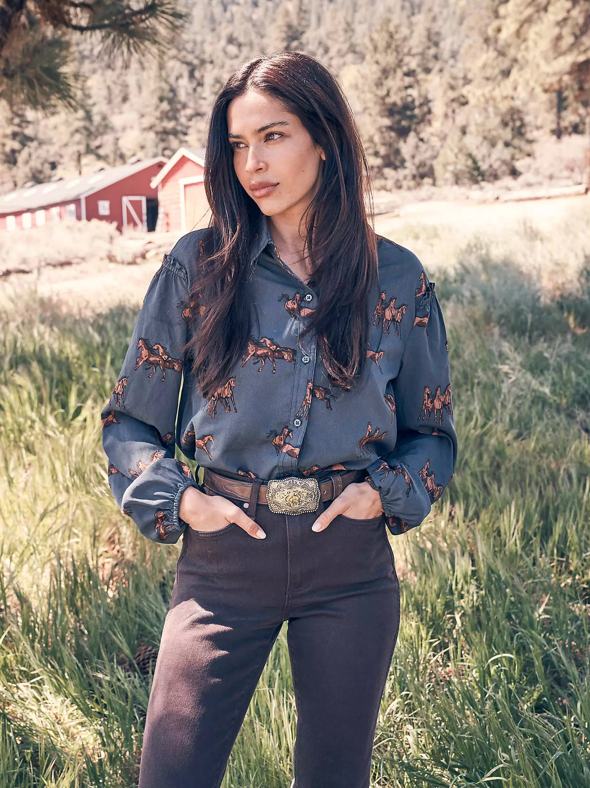 A woman standing outdoors in a grassy field wearing a long-sleeve button-up shirt with a blue-gray base and brown antler pattern, paired with dark pants and a belt.