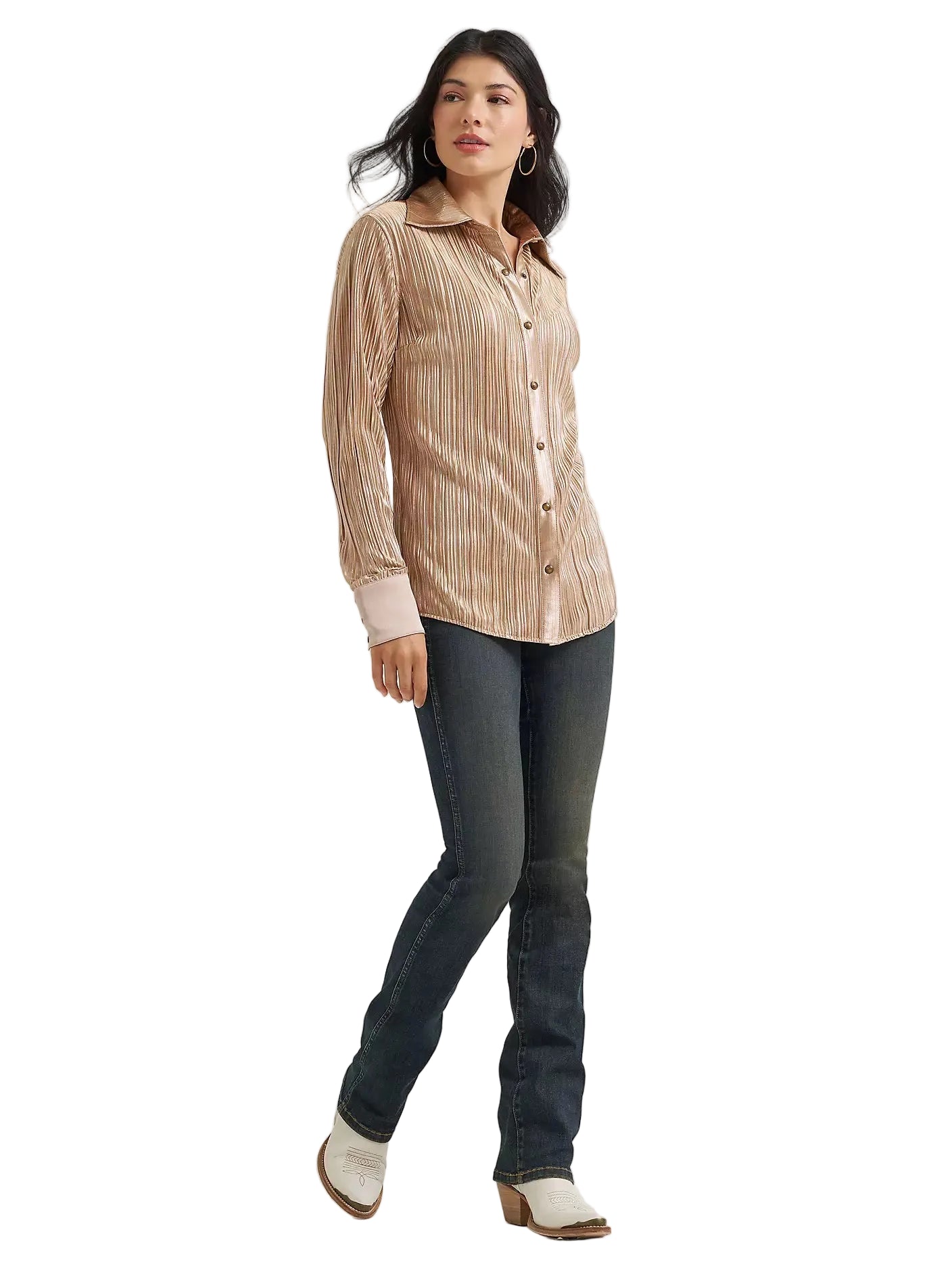 A woman modeling a long-sleeve button-up shirt with vertical pleats in a light peach-beige color, paired with dark blue jeans and white ankle boots, against a plain light gray studio background.