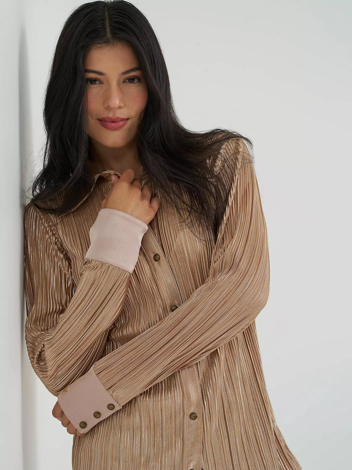 A woman wearing a pleated long-sleeve button-front top in a warm taupe-beige color with contrasting light pink cuffs, posed against a plain white background.