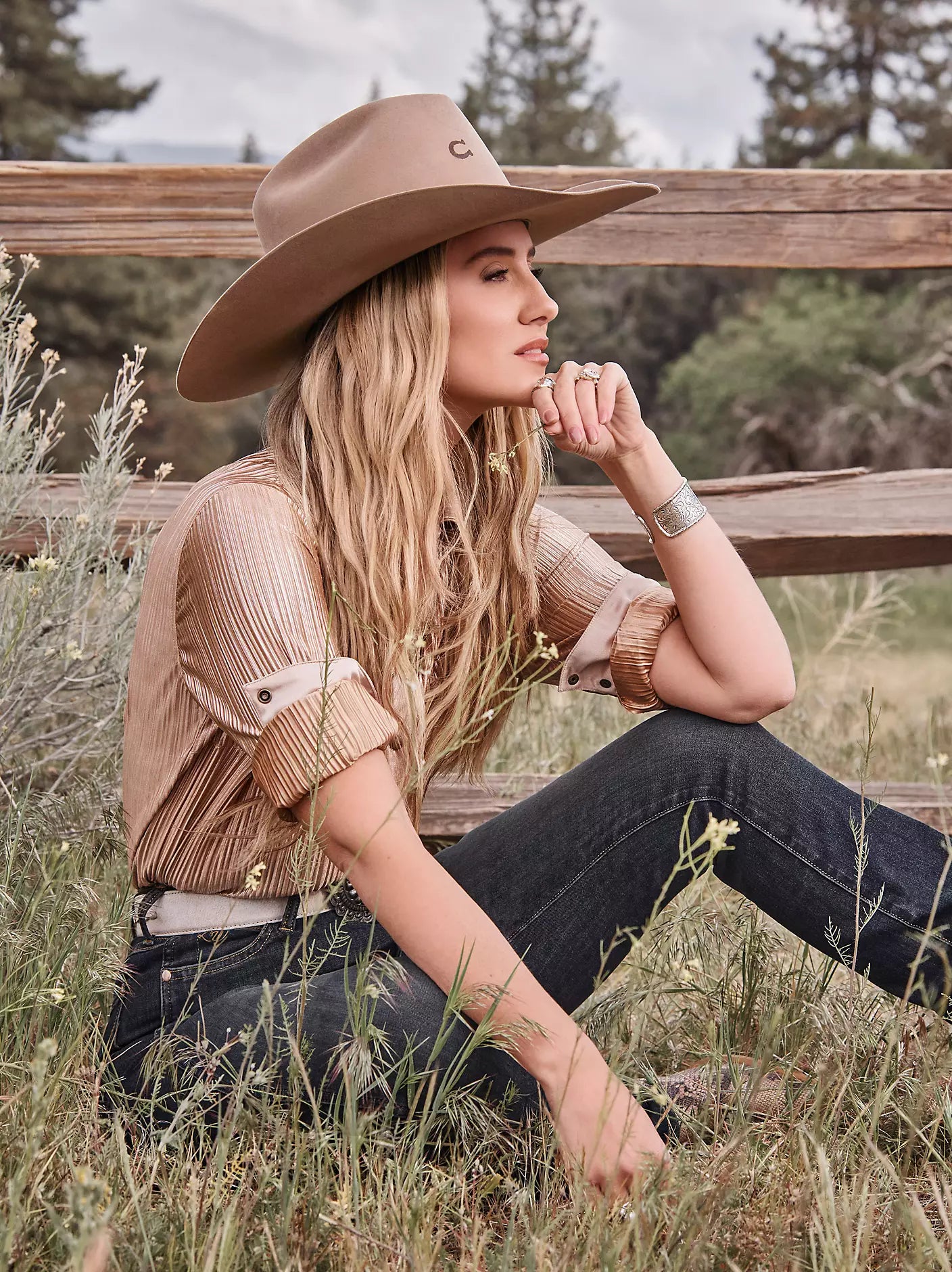 A woman sitting outdoors in a grassy field in front of a wooden fence, wearing a tan cowboy hat, a pleated tan long-sleeve shirt, and dark jeans.