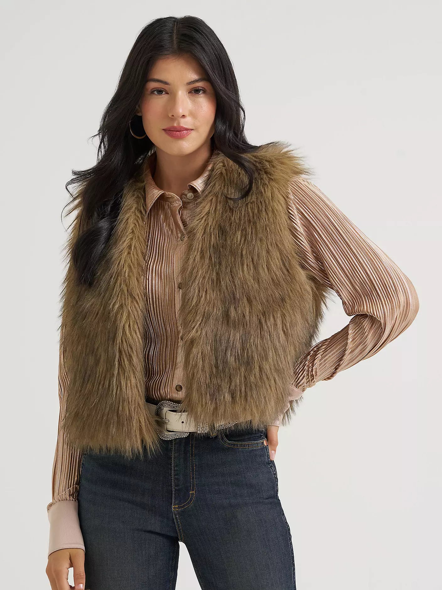 A woman wearing a tan cowboy hat with a small embroidered 'C' logo, a brown faux fur vest over a beige pleated top, and dark blue jeans, posed outdoors against a blurred forest background.