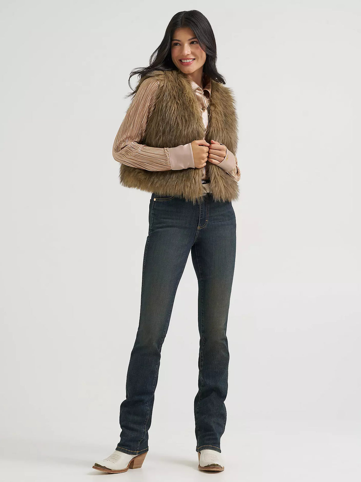 A woman modeling a cropped brown faux fur vest over a beige ribbed knit sweater, paired with dark blue jeans and white cowboy boots, against a light gray studio background.
