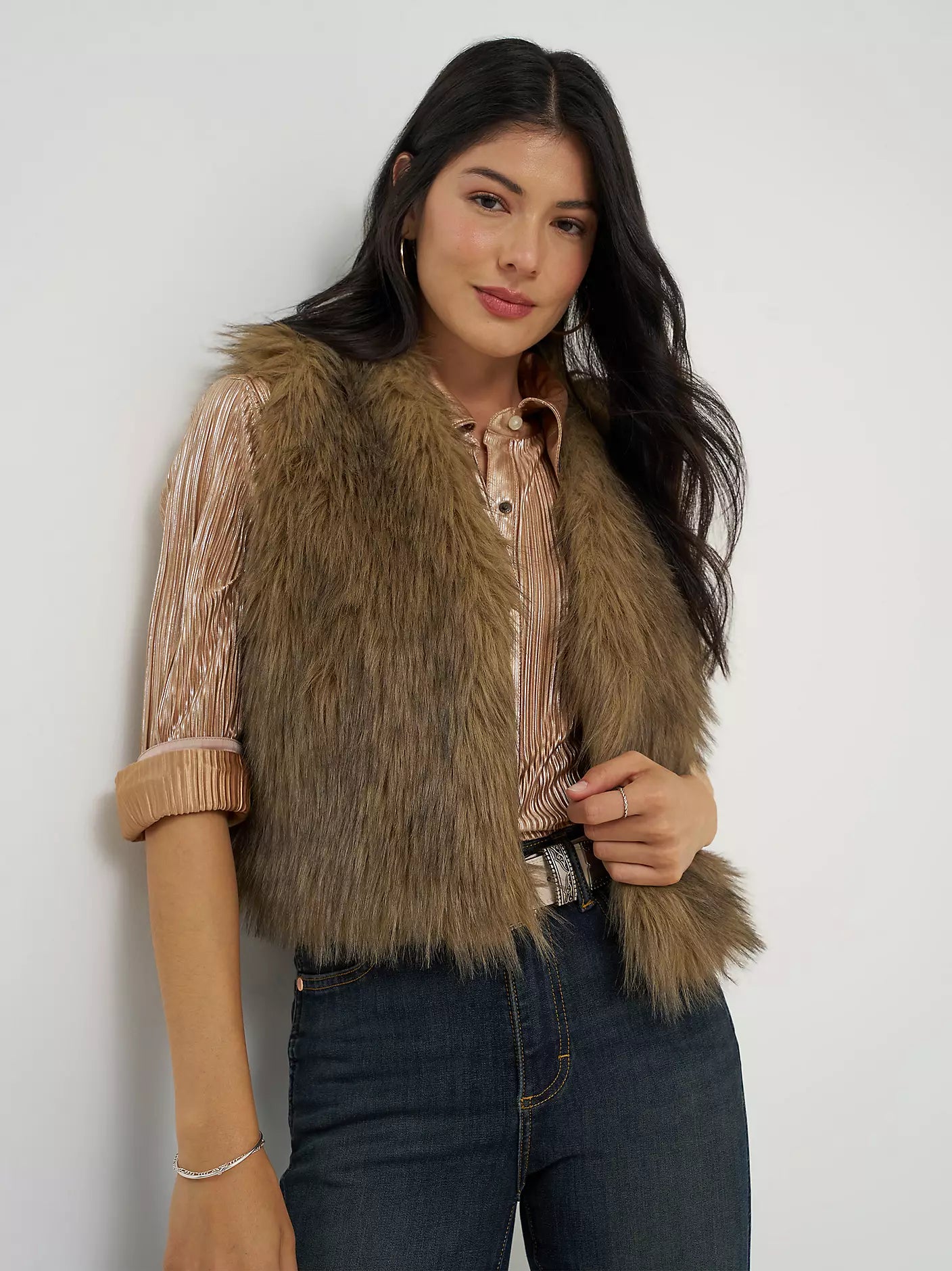 A woman wearing a brown faux fur vest over a beige striped button-up shirt and dark blue jeans, standing against a plain white wall.