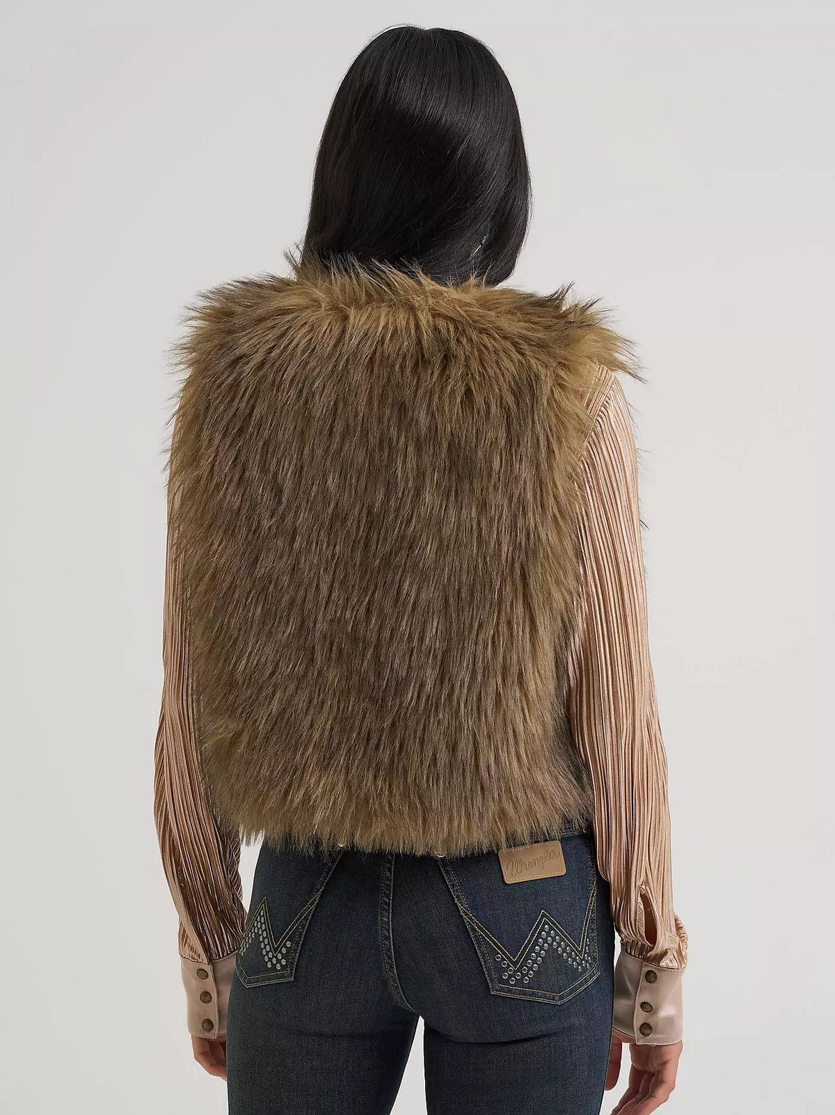 A woman shown from the back wearing a brown faux fur vest over a beige pleated long-sleeve shirt and dark blue jeans, against a plain white background.