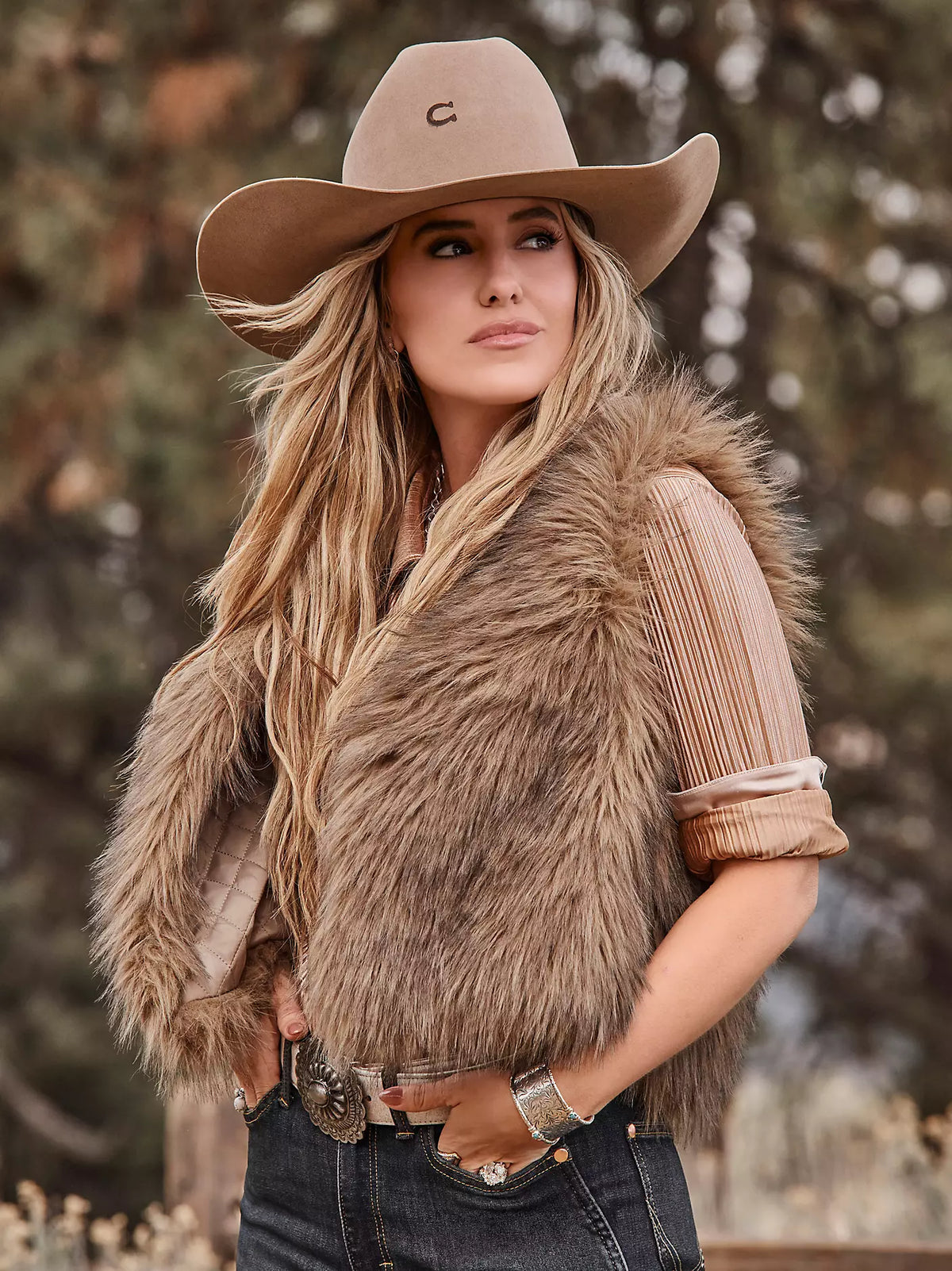 A woman wearing a tan cowboy hat with a small embroidered 'C' logo, a brown faux fur vest over a beige pleated top, and dark blue jeans, posed outdoors against a blurred forest background.