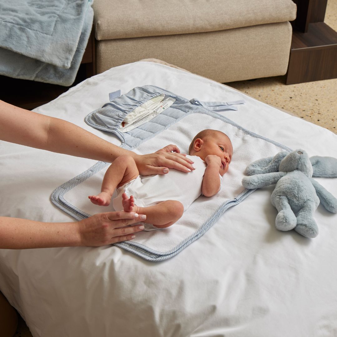 Baby lying on a white blanket with a plush toy, being held by an adult in a home setting.