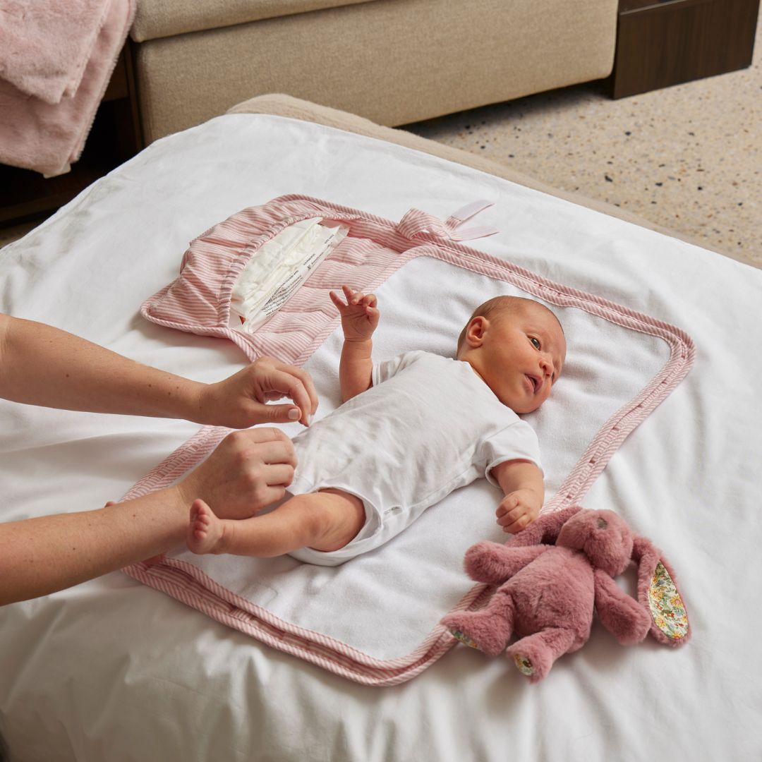 Baby lying on a white blanket with pink accents, surrounded by a plush toy and adult hands.