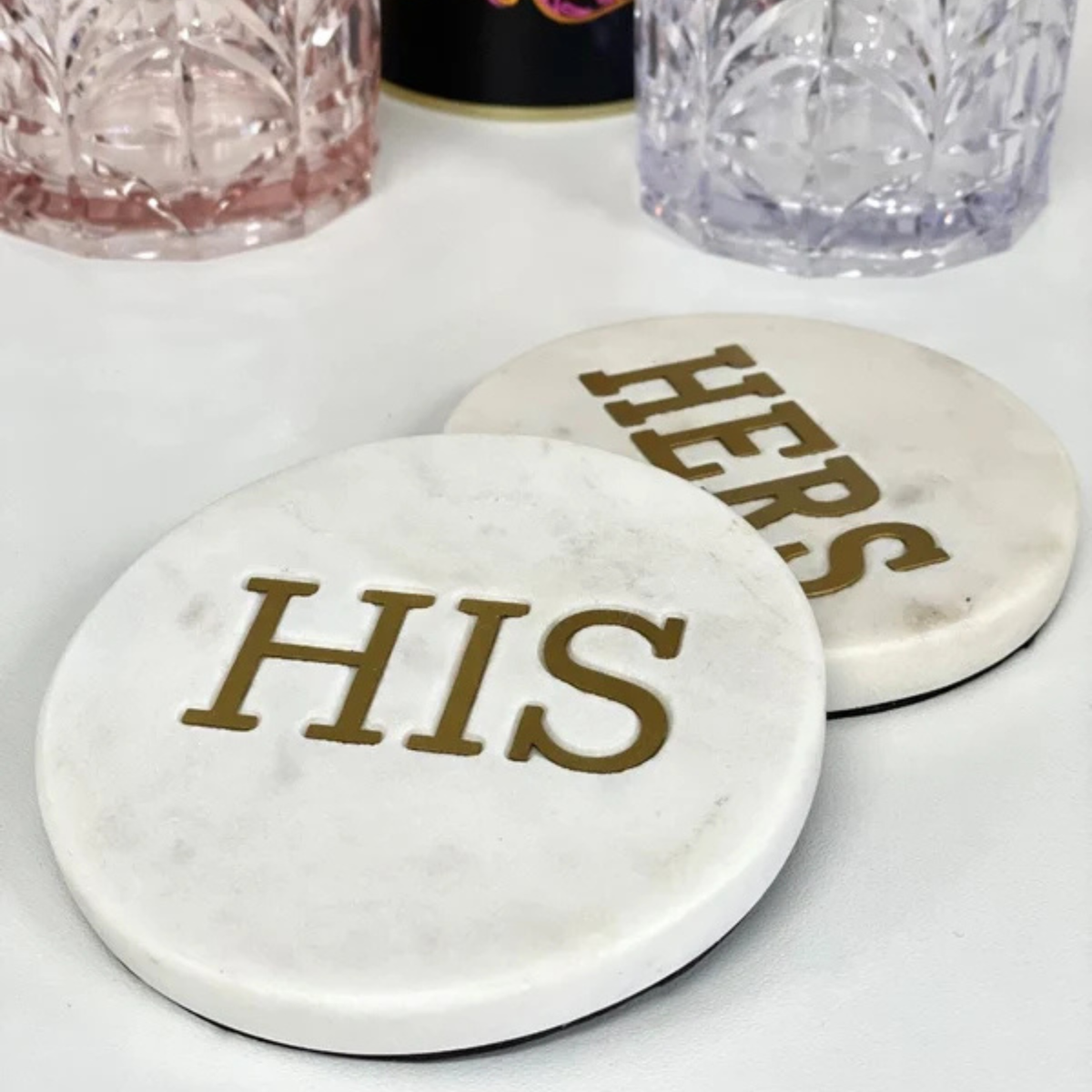 Two round white marble coasters with gold lettering, one reading 'HIS' and the other partially visible as 'HERS', shown on a white surface with decorative glasses in the background.