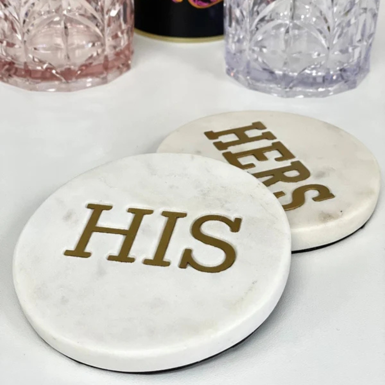Two round white marble coasters with gold lettering reading 'HERS' and 'HIS' on a white surface, with decorative glassware blurred in the background.
