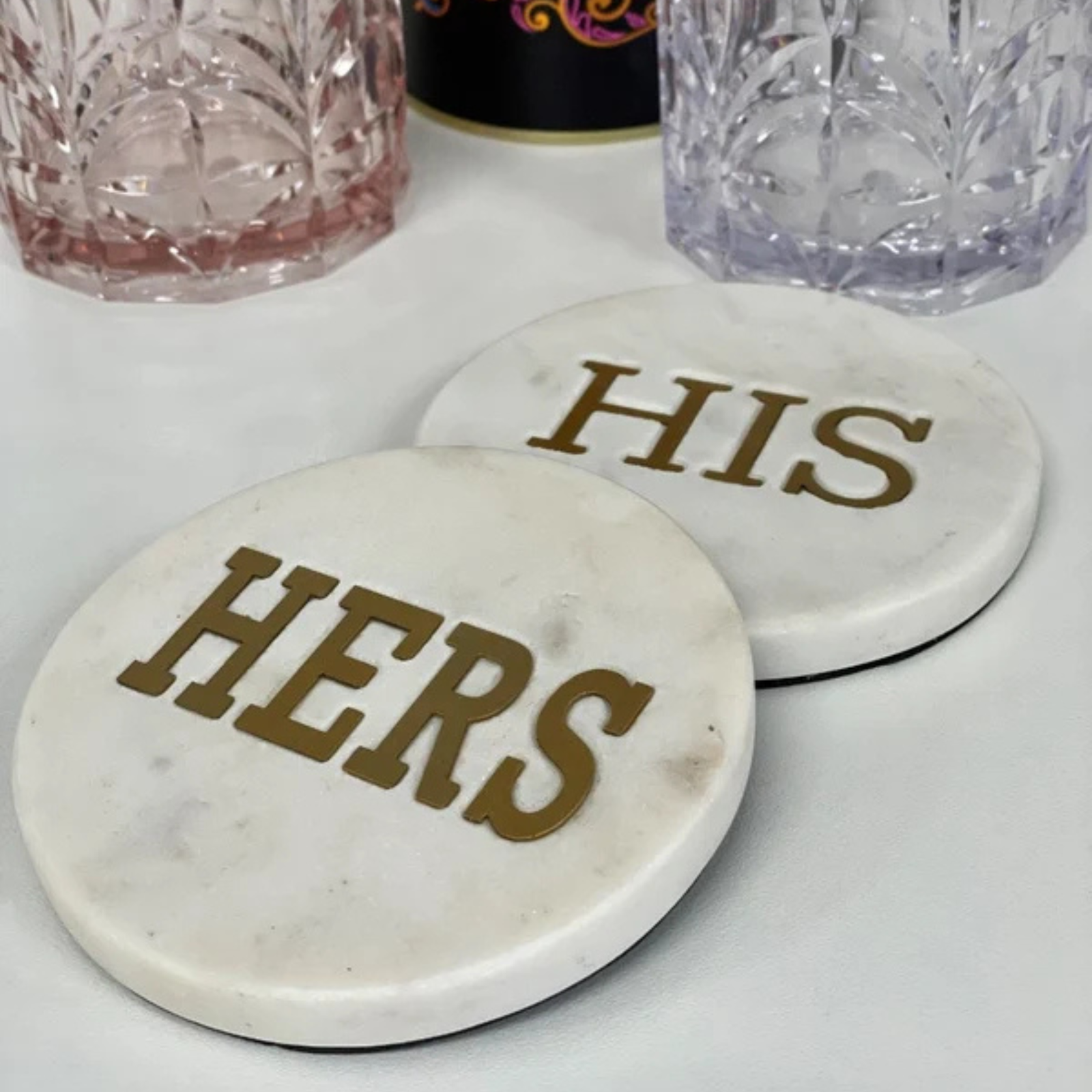 Two round white marble coasters with gold lettering reading 'HERS' and 'HIS' on a white surface, with decorative glassware blurred in the background.