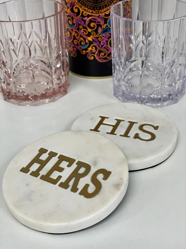 Two round white marble coasters with gold lettering reading 'HERS' and 'HIS', shown on a white surface with two faceted drinking glasses in the background.