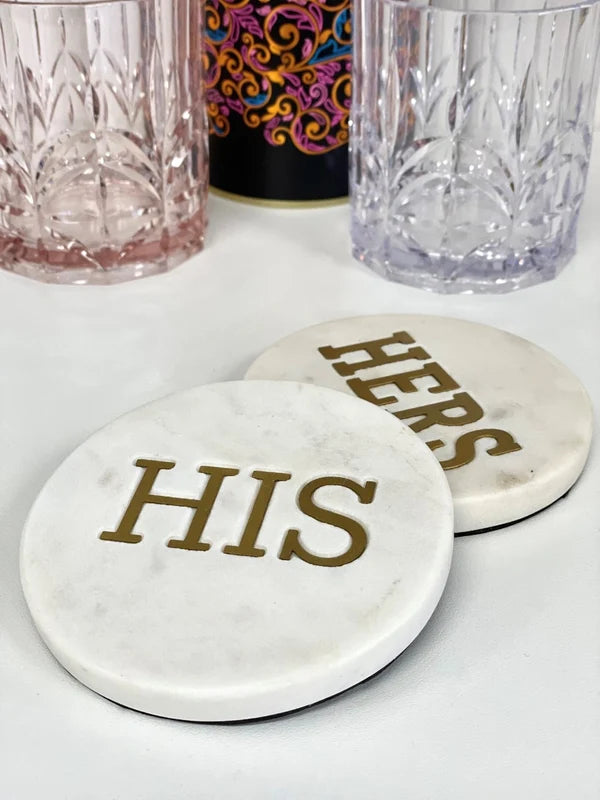 Two round white marble coasters with gold lettering reading 'HERS' and 'HIS', shown on a white surface with two faceted drinking glasses in the background.